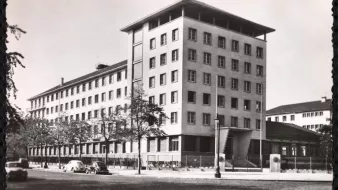 الصورة تُظهر مبنىً حديث التصميم، يتكون من عدة طوابق وله واجهة متنوعة الألوان. يتواجد حول المبنى مجموعة من الأشجار، ويبدو أن هناك شارعًا أمام المبنى حيث توجد بعض السيارات القديمة. يعكس تصميم المبنى طابعًا معماريًا بسيطًا وعصريًا، مما يوحي بأنه تم بنائه في فترة زمنية معينة في القرن العشرين. السماء تبدو واضحة مما يعطي انطباعًا بأن الصورة قد تم التقاطها في يوم مشمس.