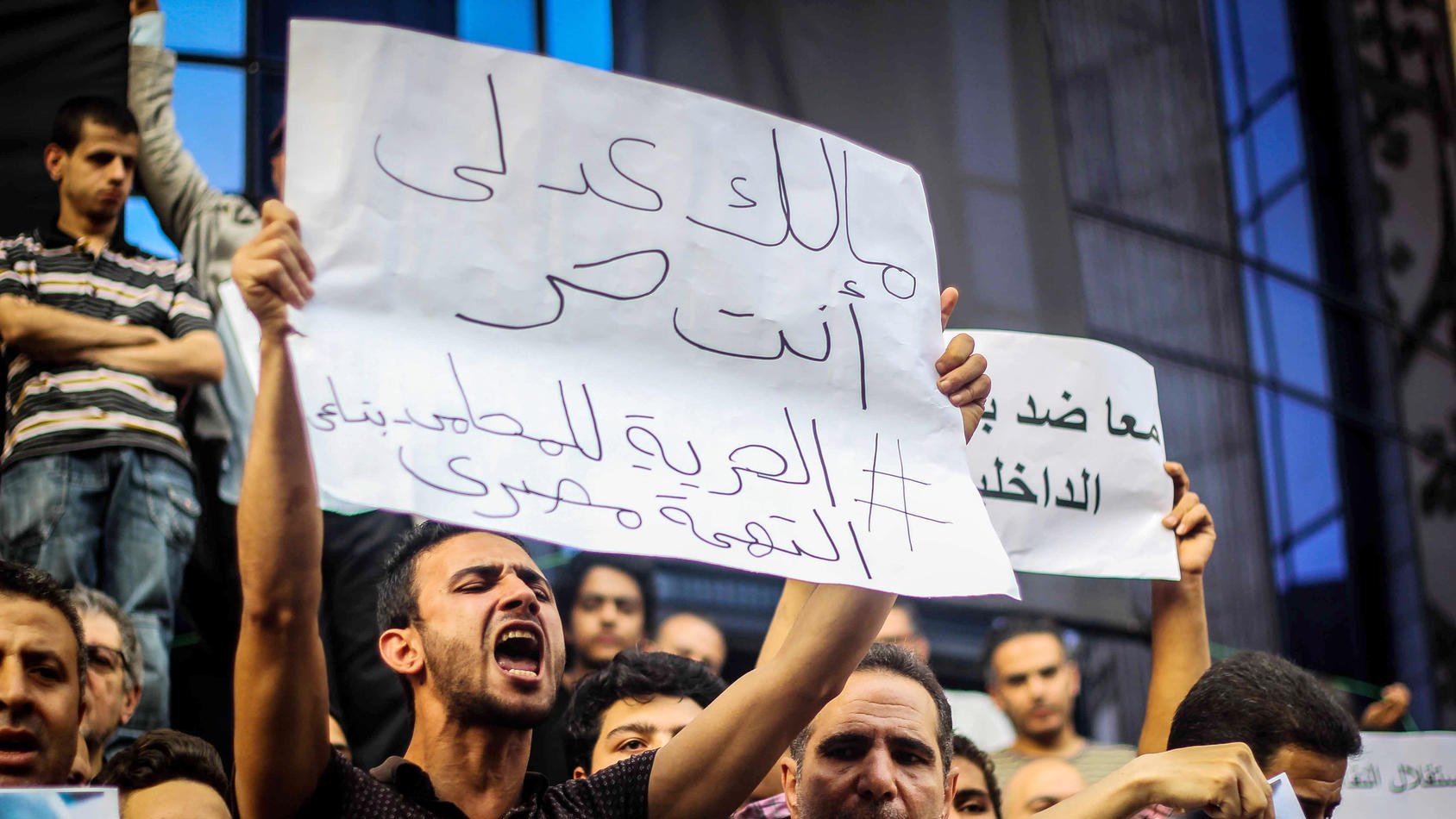 The image depicts a crowd of protestors holding signs during a demonstration. One individual is prominently featured, passionately expressing their views. The sign reads a message in Arabic, which translates to a call for freedom and justice. The crowd appears engaged and determined, highlighting the intense atmosphere of the protest. Various faces in the background show a mix of emotions, reflecting the seriousness of the situation.