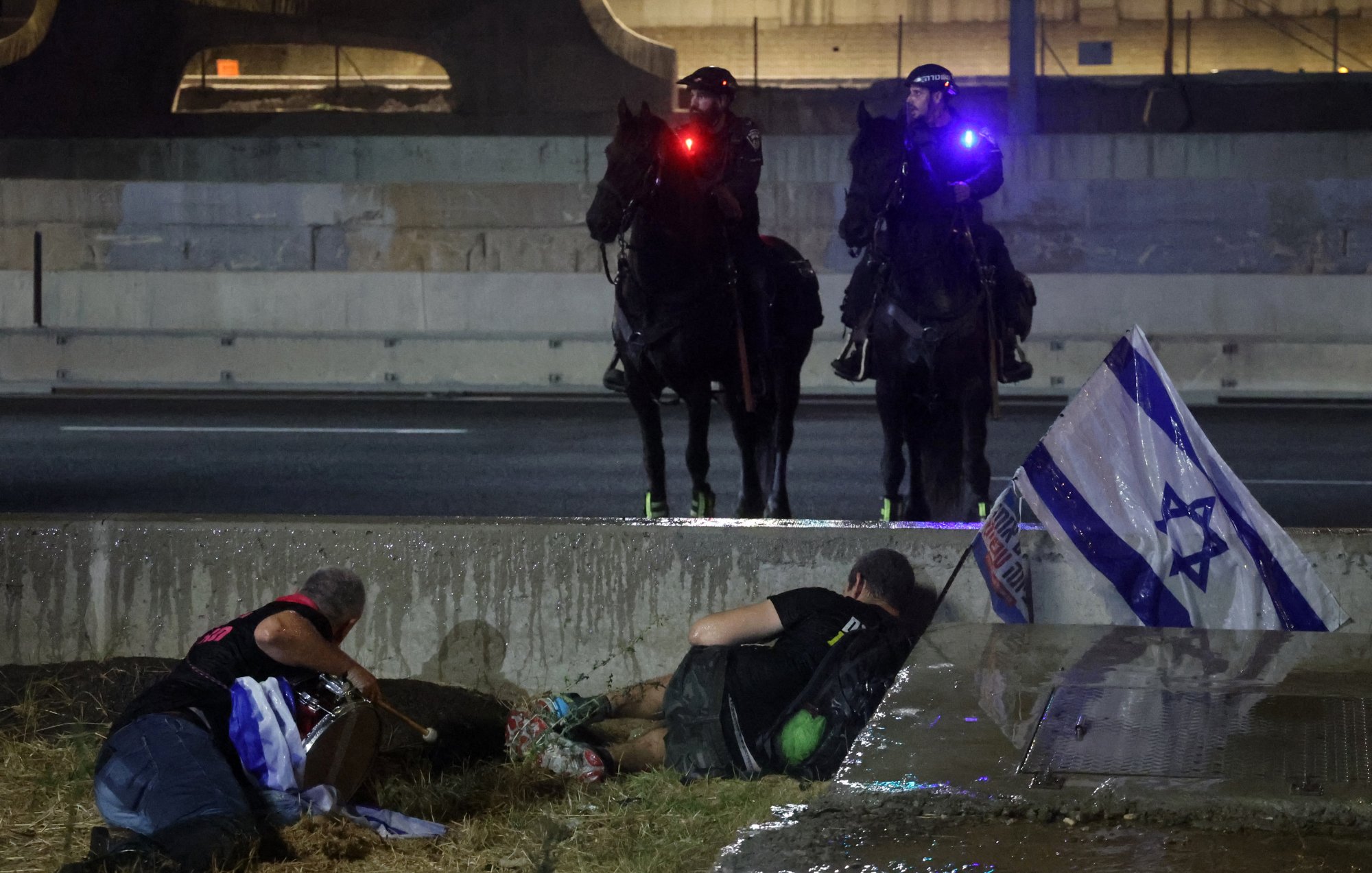 The image depicts a tense scene possibly related to a protest or civil unrest. In the foreground, two individuals appear to be on the ground, with one person lying down and another sitting nearby. They seem to be taking cover or seeking safety. In the background, two mounted police officers are visible, equipped with flashing lights on their helmets. There's also an Israeli flag positioned on the ground, suggesting that the situation might involve themes of national identity or political protest. The setting looks urban, with structures and concrete present, contributing to a sense of urgency in the atmosphere.