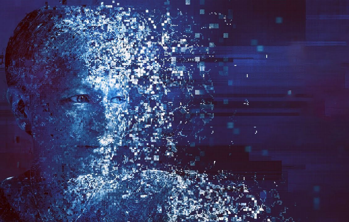 The image depicts a human-like face that appears to be disintegrating or dissolving into a myriad of small blue and white pixels or digital fragments. The background has a dark gradient with hints of purple and additional pixelated elements, creating a sense of movement and abstraction. This representation evokes themes of technology, digital transformation, and perhaps the blending of human identity with digital existence.