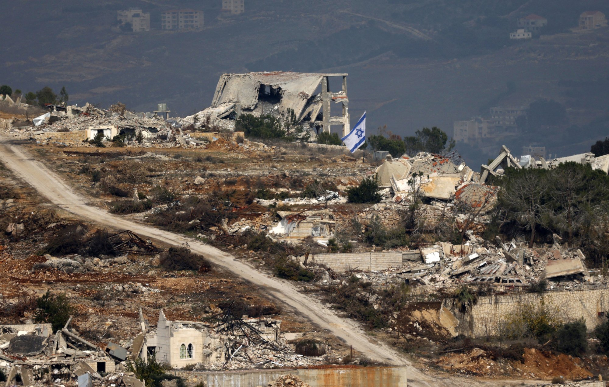 The image depicts a desolate landscape featuring extensive destruction, with several ruined buildings scattered throughout the area. In the foreground, there is a dirt road winding through the wreckage. Among the ruins, an Israeli flag can be seen prominently displayed, suggesting a connection to the location. The background shows additional structures and what appears to be a mountainous or hilly terrain, indicating a possibly rural setting. The overall scene conveys a sense of abandonment and turmoil.