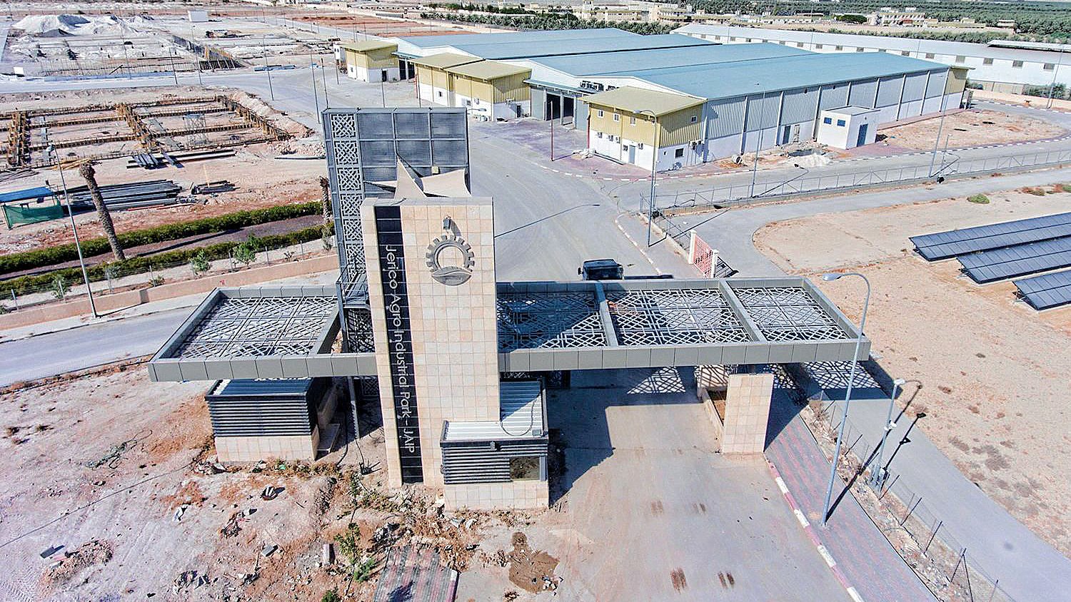 The image shows a large industrial complex with a modern entrance gate. The entrance features a prominent archway or facade, likely decorated with geometric patterns or designs. Surrounding the gate, there are several buildings, and the area appears to be part of a developing industrial site, as indicated by construction materials and equipment visible in the background. The landscape is predominantly dry and arid, with sparse greenery, and you can see a distant horizon with some smoke rising, suggesting nearby activity or construction. Solar panels are also visible on the ground, indicating a focus on sustainable energy.