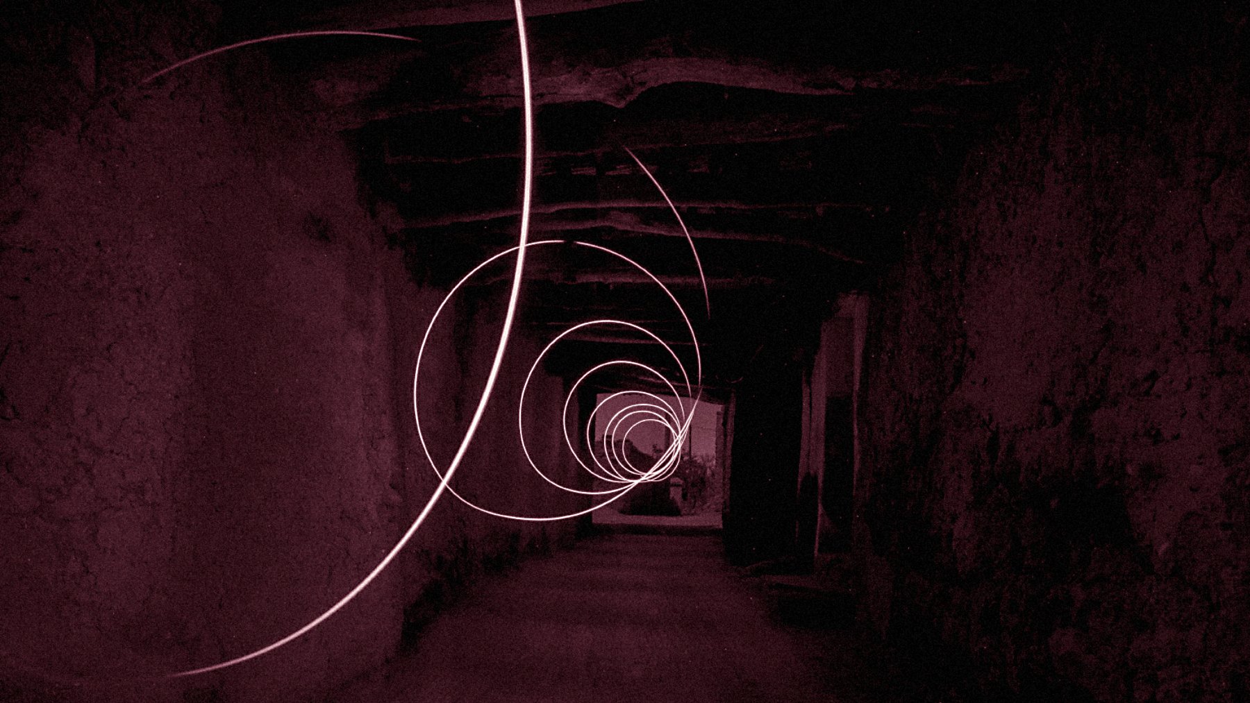 The image depicts a long, seemingly dark corridor with a series of swirling light trails creating a spiral effect. The color tone is predominantly red, contributing to a mysterious and somewhat surreal atmosphere. The walls of the corridor appear rough and textured, enhancing the sense of depth and perspective as the light trails guide the eye towards the far end of the hallway. The overall composition evokes a feeling of motion and intrigue.