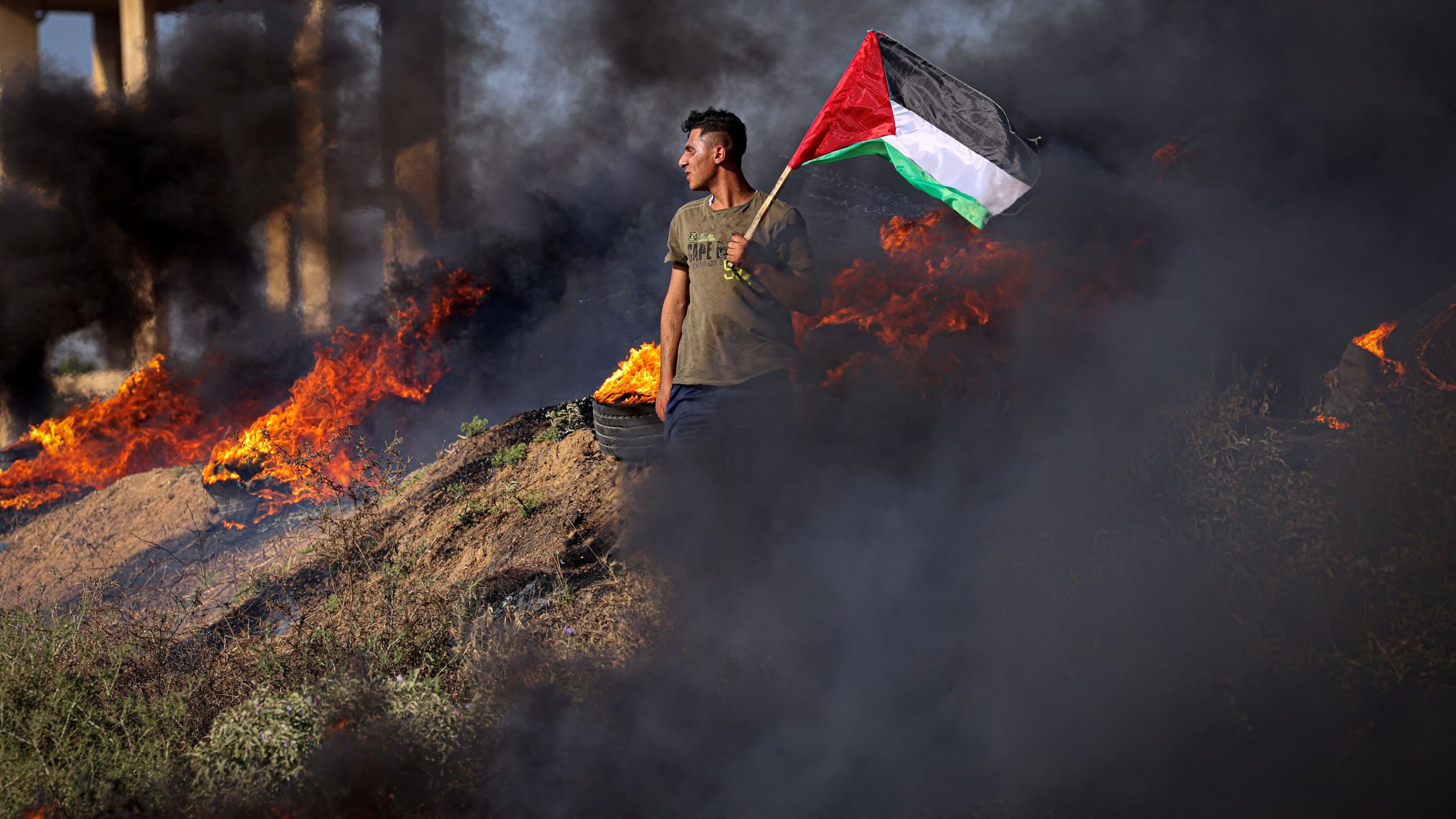 The image depicts a person standing amidst a scene of smoke and flames, holding a Palestinian flag. The individual appears to be in a protest or demonstration setting, surrounded by burning material, which creates a dramatic and intense atmosphere. The contrasting colors of the flag—red, green, black, and white—stand out against the dark smoke and fiery backdrop. The overall mood of the image conveys a sense of defiance and political expression.