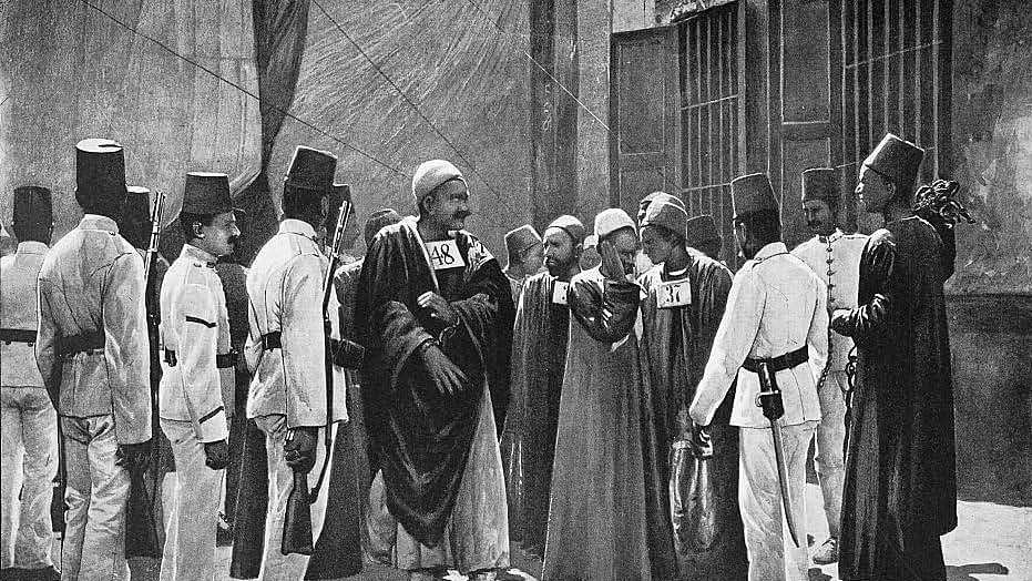 The image depicts a historical scene featuring a group of men, likely from a Middle Eastern or North African context. They are dressed in traditional attire, which includes long robes and distinctive hats. Some men are holding numbered placards, suggesting they may be part of a formal event or a historical gathering. The setting appears to be outdoors, possibly in an urban environment, with walls and structures in the background. The expressions and postures of the individuals suggest a moment of interaction or discussion among the group.