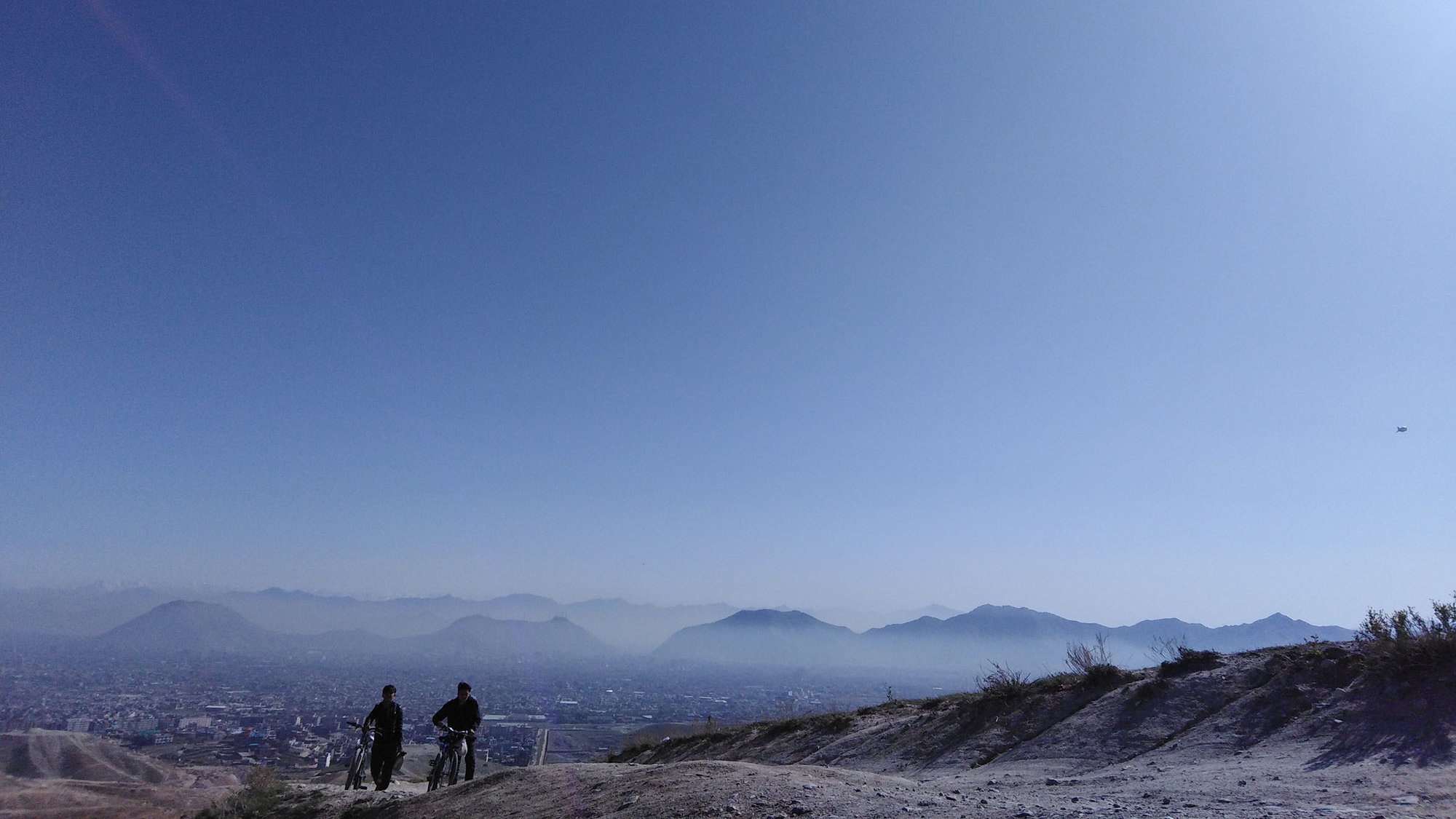 The image depicts a serene landscape with two individuals walking along a trail on a hillside. They are silhouetted against a vast, clear blue sky, which suggests a sunny day. In the background, there are distant mountains and a sprawling cityscape, partially visible below. The scene captures a sense of tranquility and openness, highlighting the natural beauty of the setting.