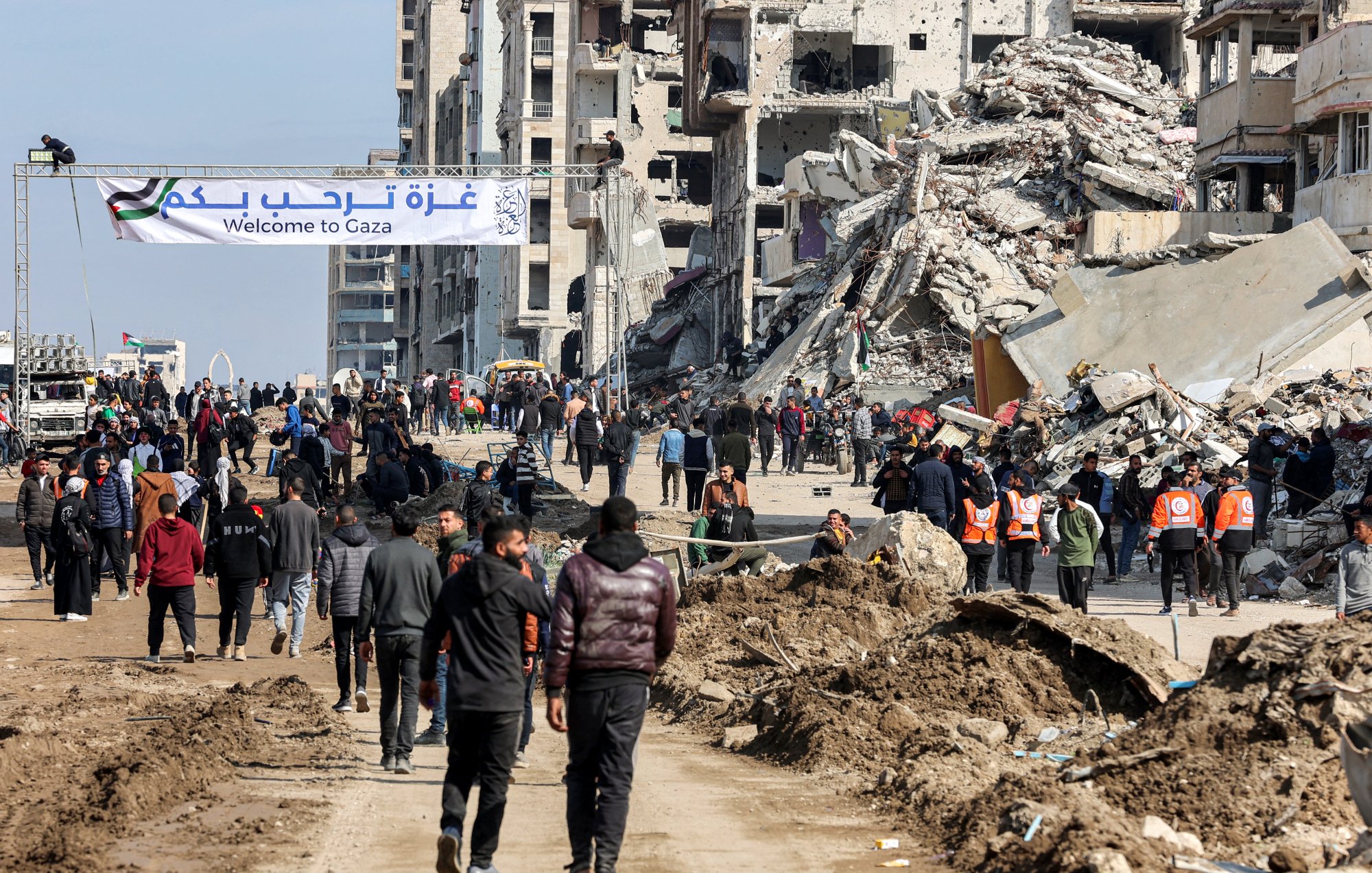 The image depicts a scene in Gaza featuring a street filled with people walking. In the background, there are buildings that appear to be damaged or destroyed. A large banner stretches across the street, welcoming visitors to Gaza, and some individuals wearing orange vests are visible, likely indicating they are part of a relief or aid organization. The area is marked by piles of debris and dirt, suggesting recent conflict or disaster. Overall, the atmosphere conveys a mix of resilience amid the challenges faced by the community.