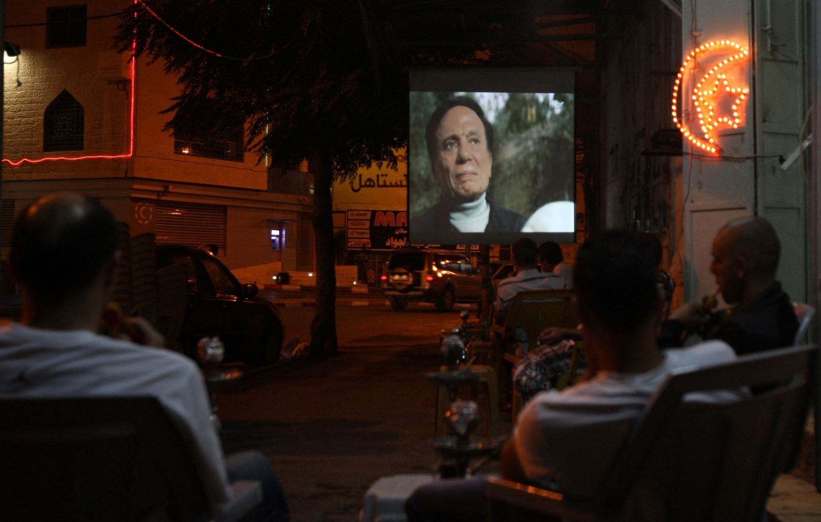 The image depicts a night scene where a group of people is seated in chairs, watching a film projected onto a large screen set up outside. The atmosphere appears relaxed, with some of the viewers enjoying drinks, possibly shisha. The area is illuminated by both the projection light and neon signage in the background, adding a lively ambiance to the setting. The scene captures a communal experience centered around outdoor movie viewing.