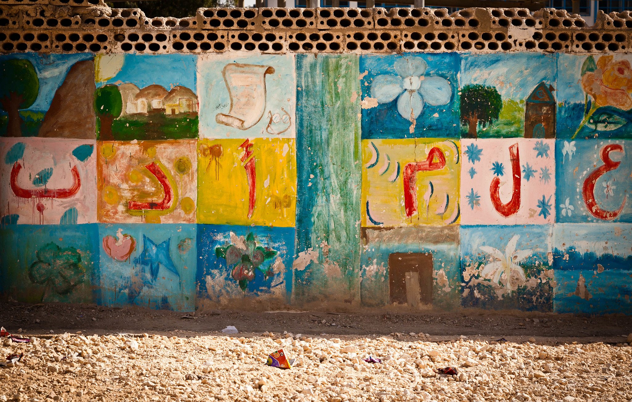 The image shows a wall decorated with colorful murals or paintings. The artwork includes various designs such as flowers, shapes, and possibly symbols or letters that are arranged in a grid pattern. The colors are vibrant, featuring shades of blue, green, yellow, and pink, giving the wall a lively and artistic feel. The surrounding area appears dusty or sandy, suggesting an outdoor environment. Overall, the scene conveys a sense of creativity and community.