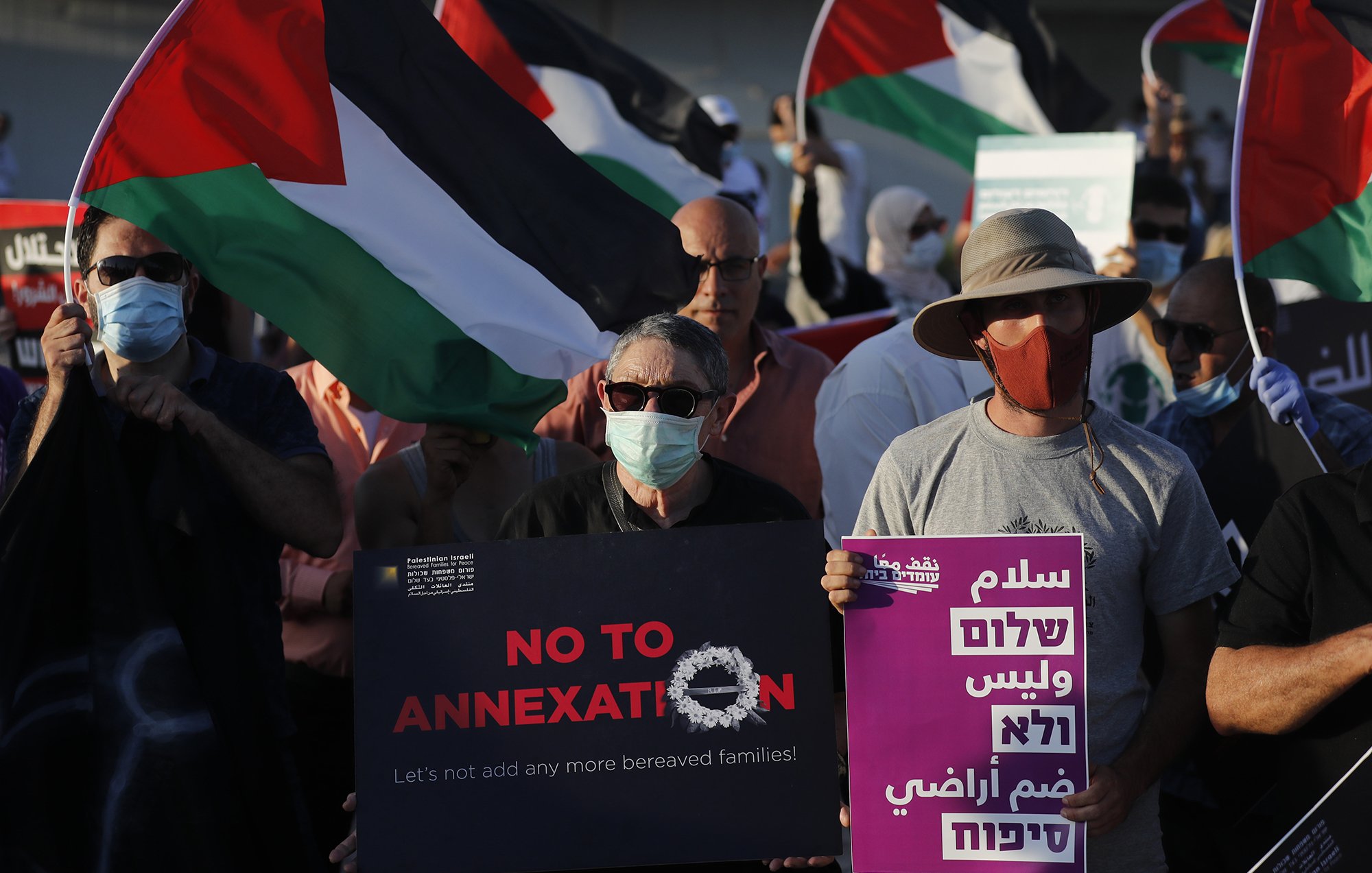 The image depicts a group of protesters holding flags and signs during a demonstration. The flags feature the colors of the Palestinian flag. Some participants are wearing masks, and a few signs read messages against annexation and calls for peace. The atmosphere appears to be one of solidarity and activism, as the individuals gather for a common cause.