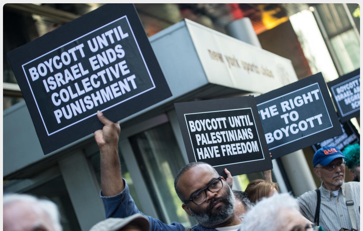 The image depicts a group of protesters holding signs at a demonstration. Some of the signs read "BOYCOTT UNTIL ISRAEL ENDS COLLECTIVE PUNISHMENT" and "BOYCOTT UNTIL PALESTINIANS HAVE FREEDOM." The atmosphere suggests a call for social justice and awareness regarding the Palestinian situation. The protesters appear engaged and determined in their stance.