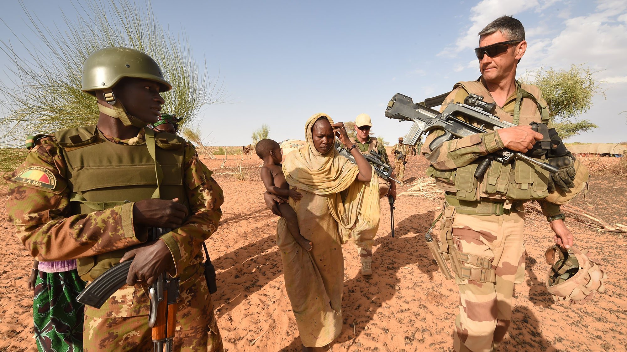 The image shows a scene in a desert setting, featuring military personnel and civilians. On the left, a soldier in a camouflage uniform holds an automatic weapon and wears a helmet, while next to him, a woman in traditional attire carries a young child. To the right, another soldier, dressed in a different military uniform and also armed, is observing the surroundings. The background features sparse vegetation typical of a desert environment. The overall atmosphere suggests a military or peacekeeping operation, possibly in a humanitarian context.