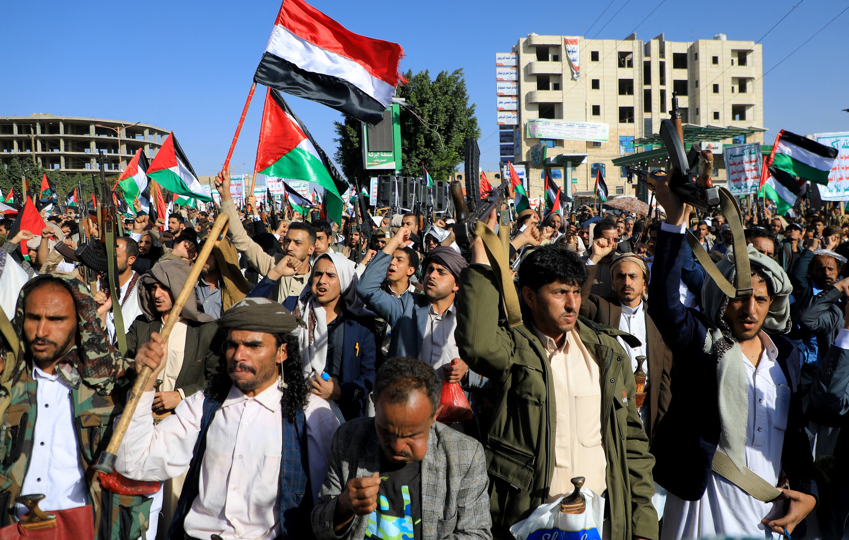 The image depicts a large crowd of people gathered in an outdoor setting, likely during a protest or rally. Many individuals are holding flags, including ones featuring a red, white, and black design, which suggests a political or national theme. The crowd appears energized, with some participants raising their fists or flags. In the background, there are buildings, indicating an urban environment. Overall, the scene conveys a strong sense of collective action and expression.