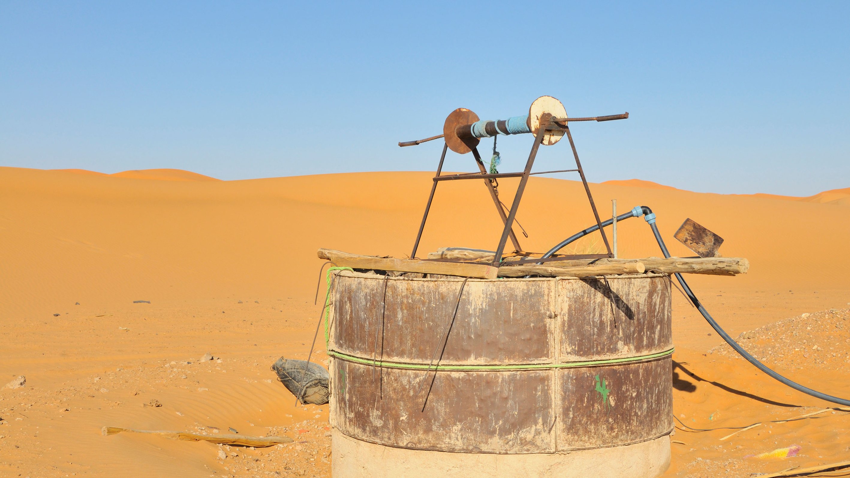 The image shows an old water well situated in a vast desert landscape. The well is made from a large, cylindrical structure with a rusty metal exterior and a concrete base. At the top, there is a simple wooden frame holding a pulley and a couple of wheels, likely used for drawing water. Surrounding the well are sand dunes, displaying a warm golden color under a clear blue sky, emphasizing the arid environment.