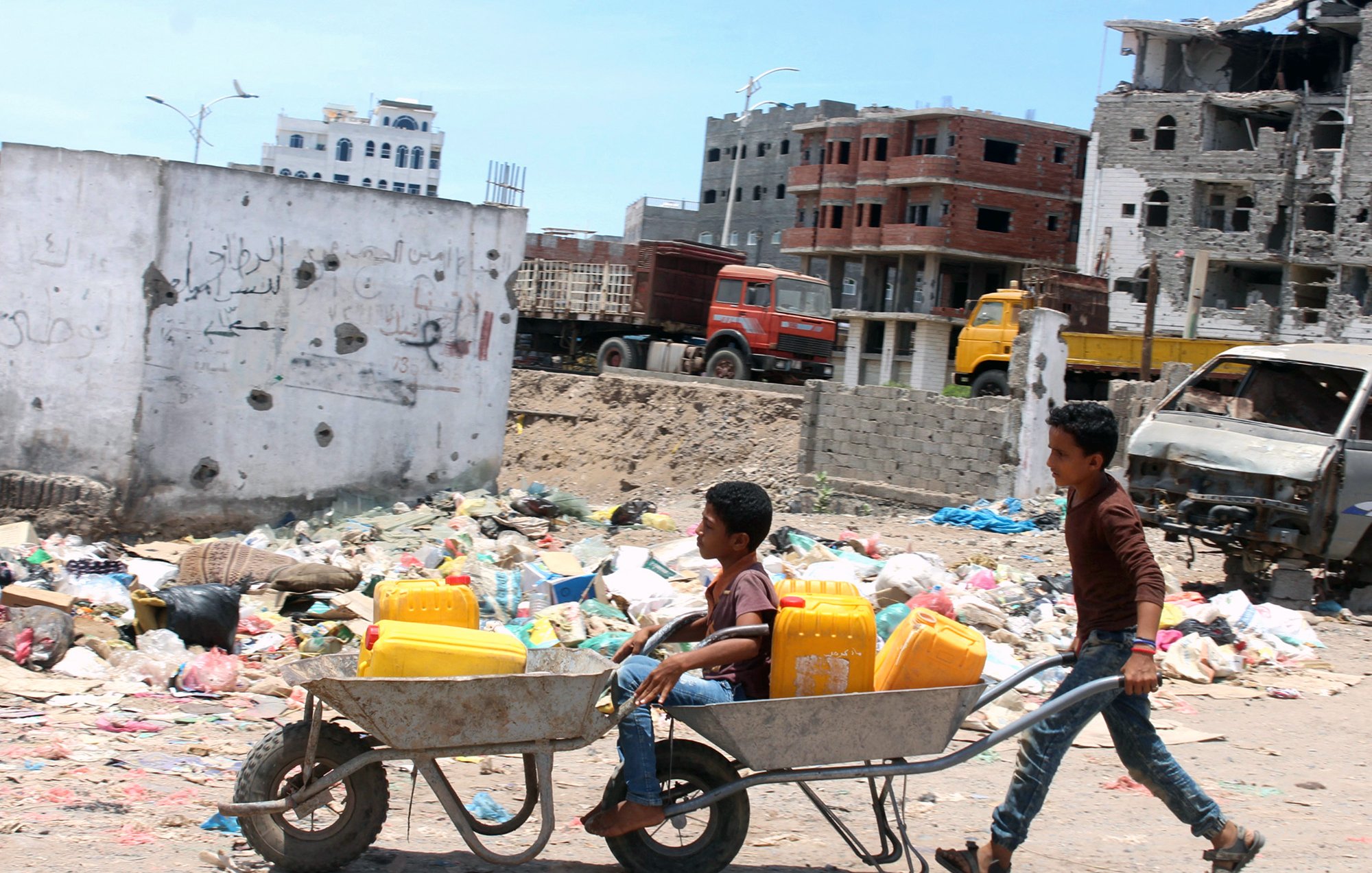 The image shows two boys in a dilapidated urban setting. One boy is pushing a wheelbarrow that carries another boy sitting inside it. The background includes a scene of garbage and rubble, with partially constructed buildings and damaged structures. The environment appears to be neglected, highlighting issues related to poverty and urban decay. The sky is clear, suggesting a bright day.
