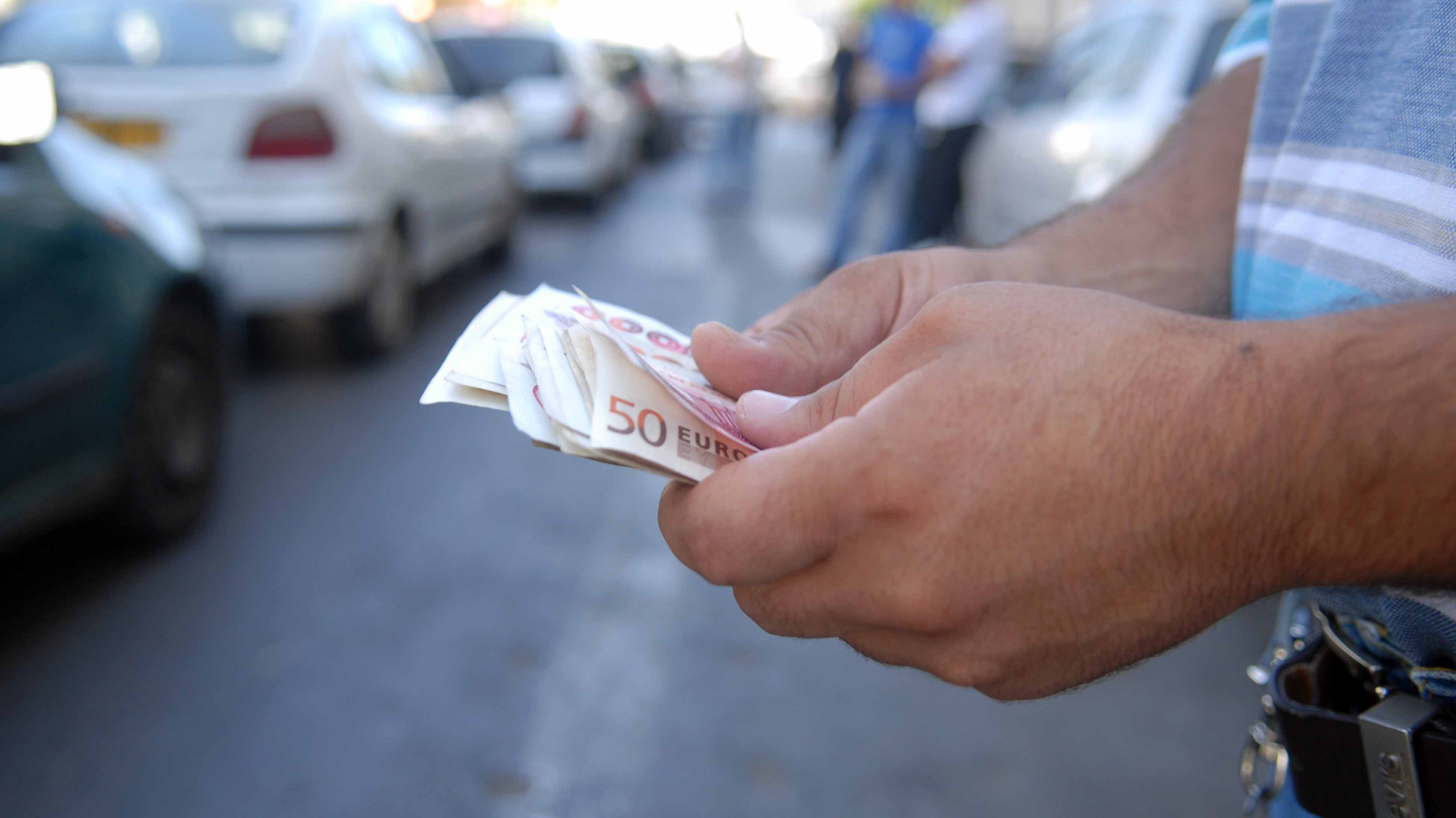 The image shows a person's hands holding several folded banknotes, specifically a stack of 50-euro notes. In the background, you can see vehicles parked along a street, indicating an urban setting. The focus is on the cash being counted or examined, suggesting a financial transaction or a moment of handling money.