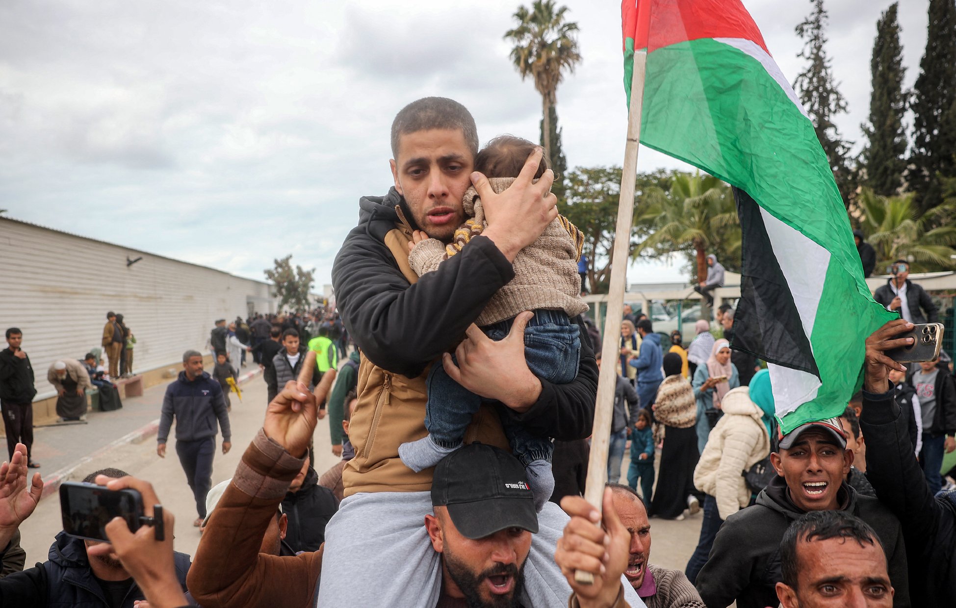 The image depicts a scene of a gathering or demonstration. A man is holding a child closely to him, showcasing a protective gesture. Around them, a crowd appears to be rallying, with some individuals raising their hands and a prominent Palestinian flag visible in the background. The atmosphere seems charged, indicating a moment of significance, possibly related to a political or social issue. The setting includes trees and a building in the background, against a cloudy sky.