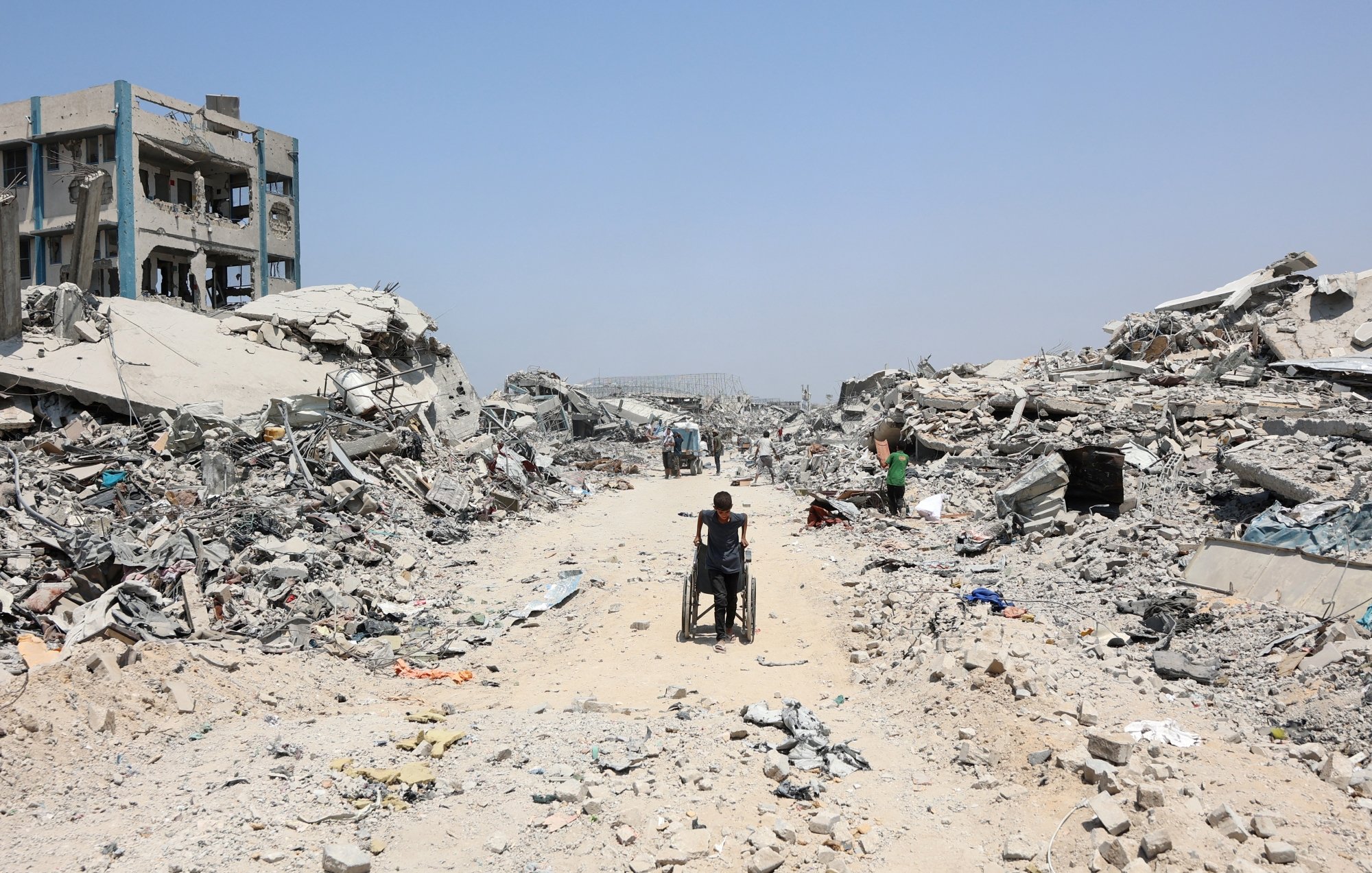 A person in a wheelchair navigates a debris-filled street amid collapsed buildings.