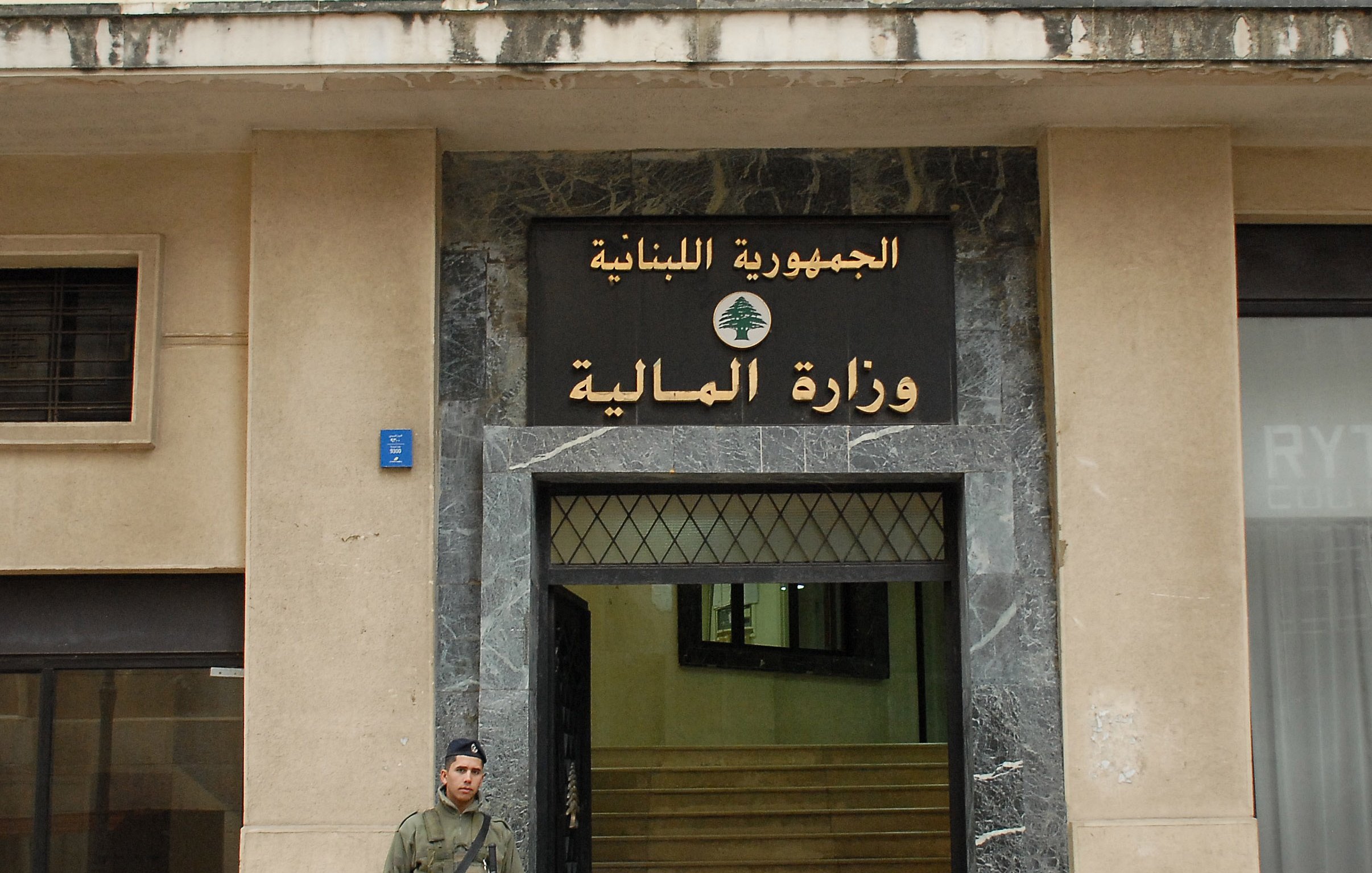 The image shows a soldier standing outside a building with a large sign displaying the words "الجمهورية اللبنانية" (Lebanese Republic) and "وزارة المالية" (Ministry of Finance) in Arabic. The building has a modern façade with a marble entrance. The soldier is wearing military uniform and holding a weapon, indicating a security presence at the site. The overall setting suggests a government location in Lebanon.