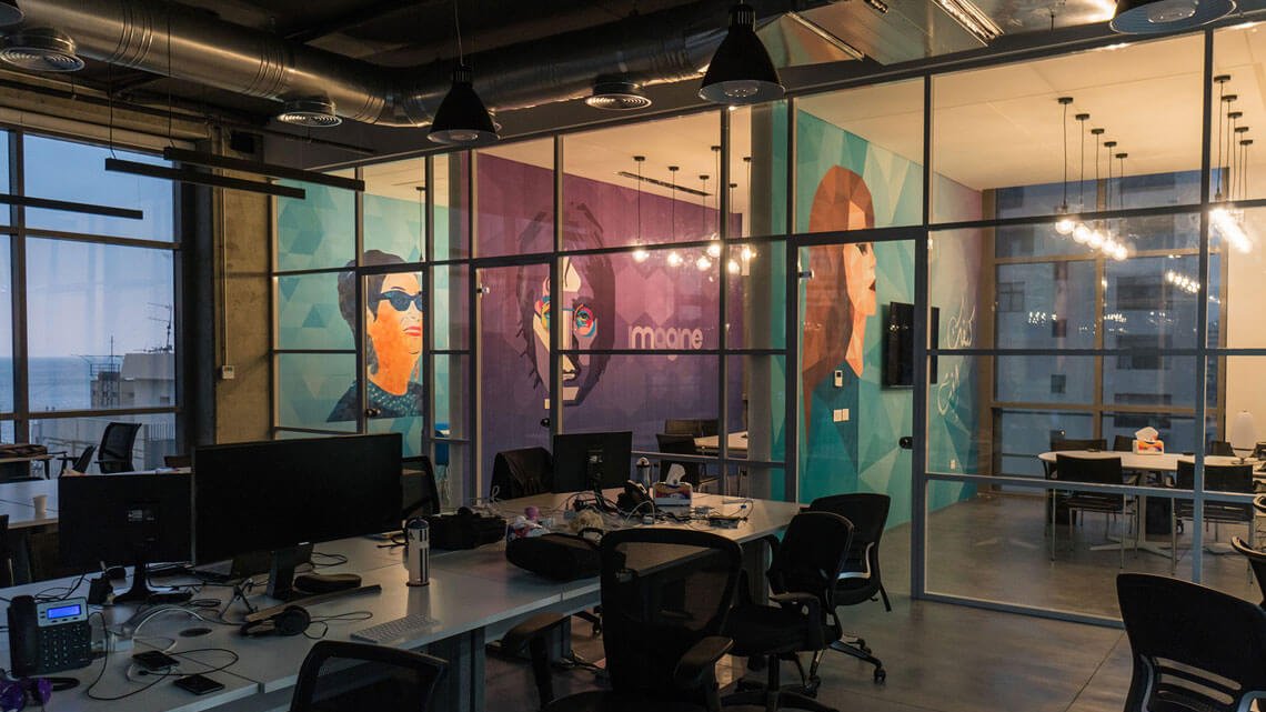 The image shows a modern office space with large glass windows, providing a view of the outside. Inside, there are several workstations with computers and ergonomic chairs. The walls are decorated with vibrant murals featuring stylized portraits of individuals, adding a creative touch to the environment. The overall ambiance is open and contemporary, with industrial elements like exposed ducts and warm lighting fixtures. The space appears to be designed for collaboration and innovation.
