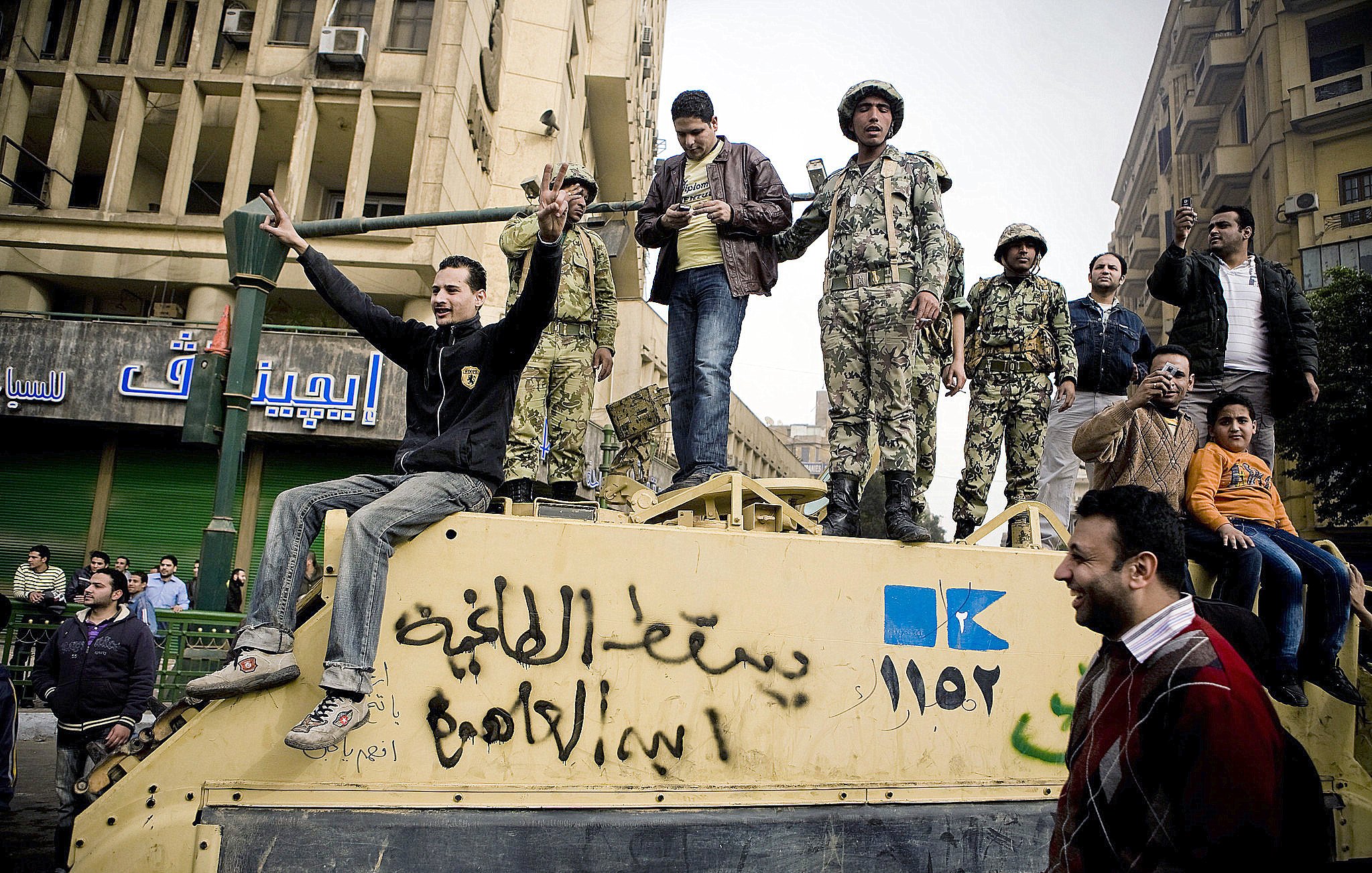 The image depicts a group of people, including soldiers, gathered on top of a military vehicle in an urban setting. Some individuals are gesturing and expressing emotions, while others appear to be observing or interacting with the crowd below. The environment is bustling, with buildings visible in the background, indicating a scene of protest or public demonstration. The atmosphere suggests a moment of tension or significant political activity.