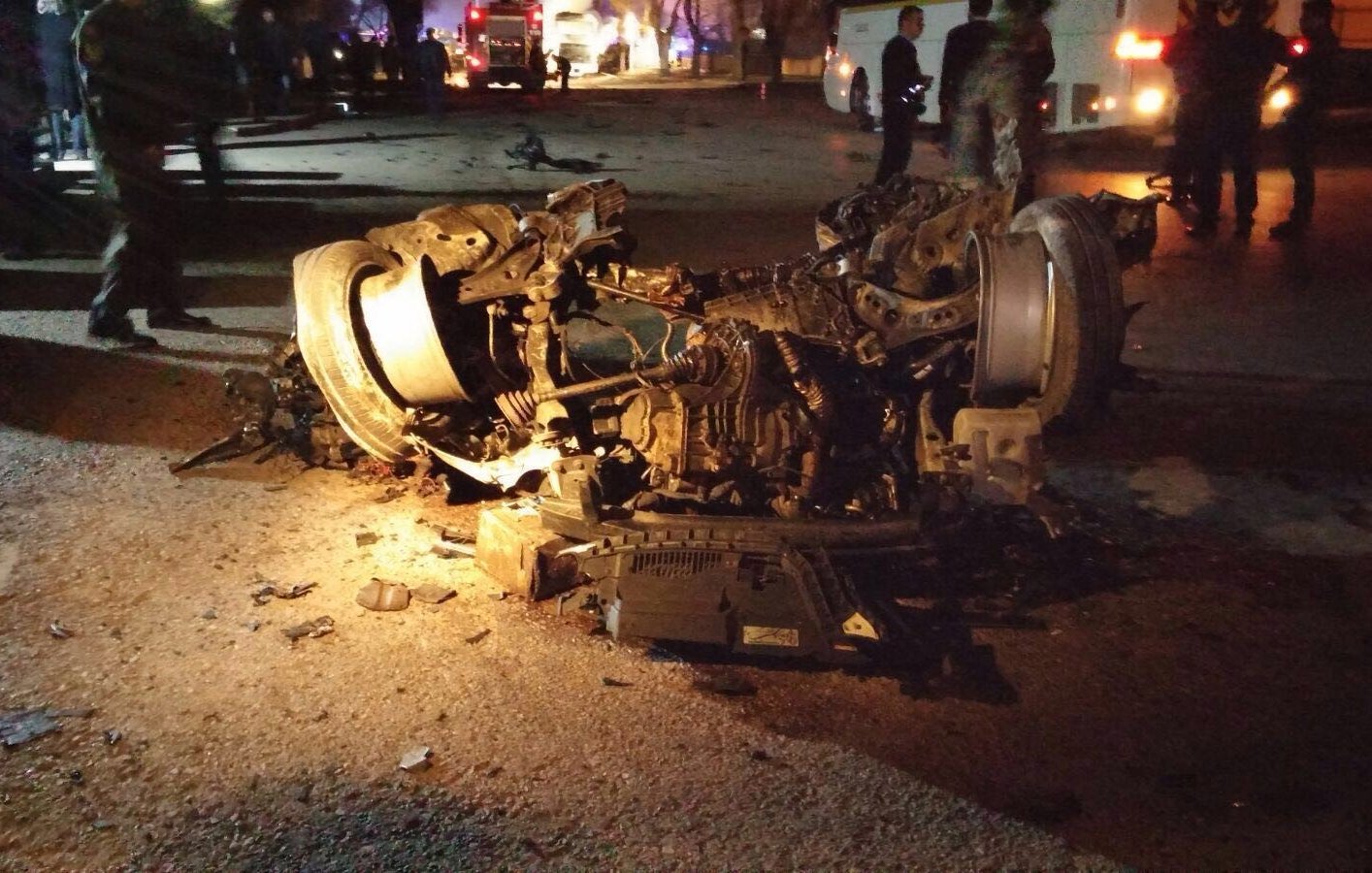 The image depicts a severely damaged vehicle that has been overturned. The car's chassis and engine are exposed, showing significant destruction, with debris scattered around the area. The scene appears to be dark, possibly at night, with some emergency responders or bystanders in the background, indicating that it may have been the site of an accident. The overall context suggests a serious incident involving the vehicle.