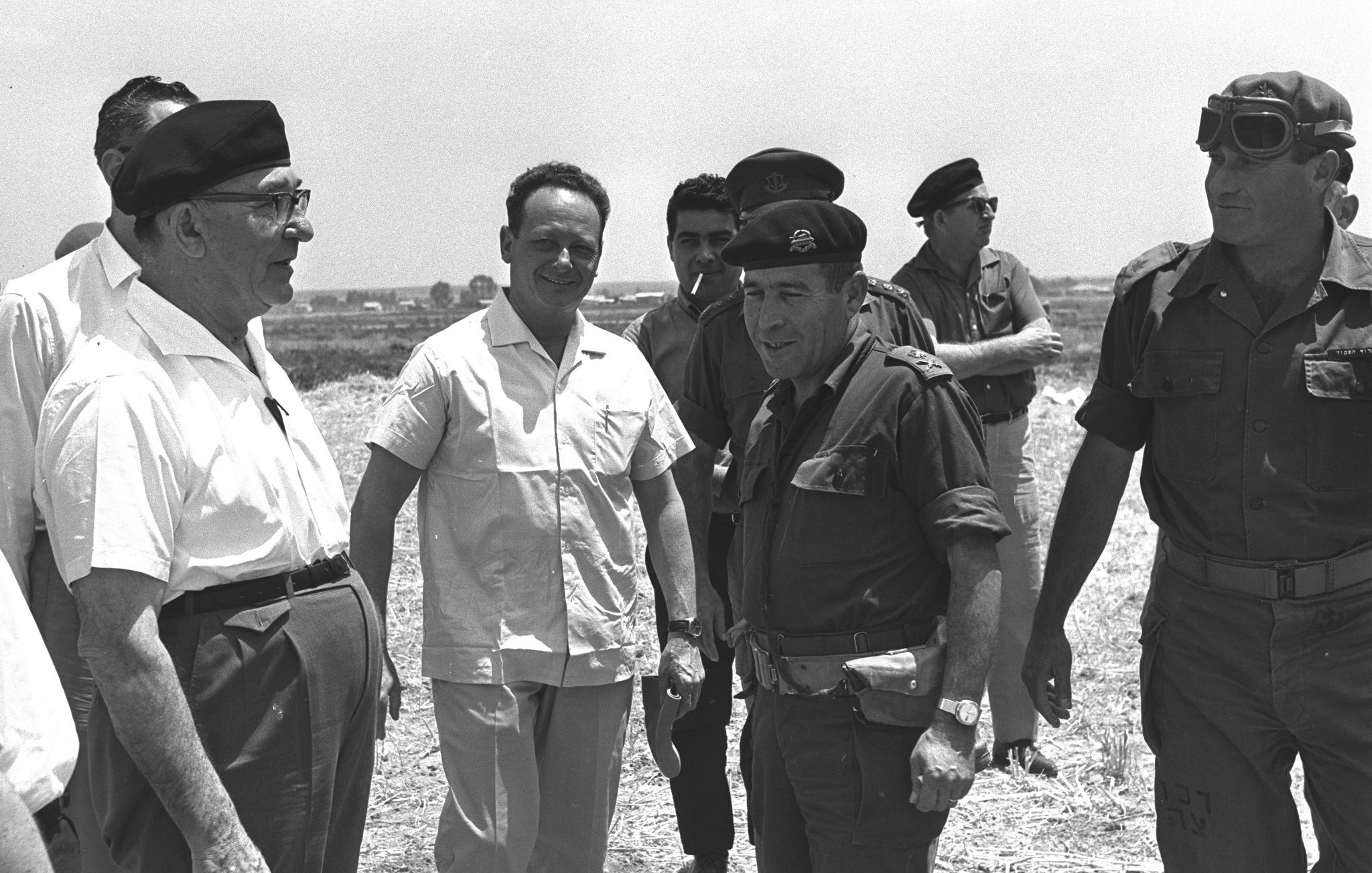 The image appears to show a group of men in a casual outdoor setting, likely involved in a military or political context. Some individuals are dressed in military uniforms, while others wear casual or formal attire. The atmosphere seems informal as they engage in conversation, suggesting a moment of collaboration or discussion. The background features a rural landscape, possibly indicating a visit to a site of significance.