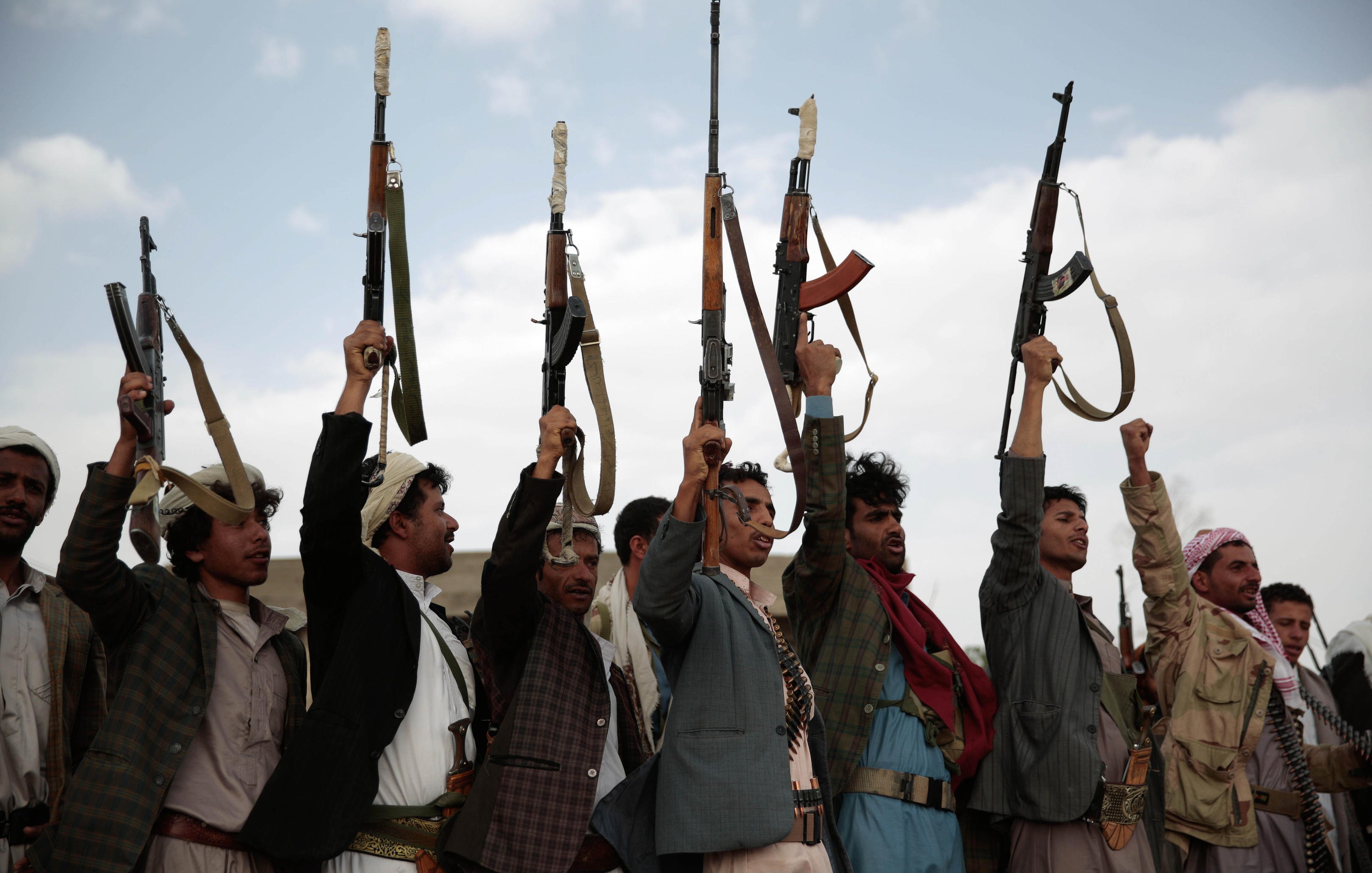 The image shows a group of men holding up rifles in the air. They are standing in a line, with some raising their right arms in a fist while others are focusing on the guns. The setting appears to be outdoors, with a cloudy sky in the background. The men are wearing traditional attire, which varies among them, and there is an overall sense of solidarity and determination in their postures.