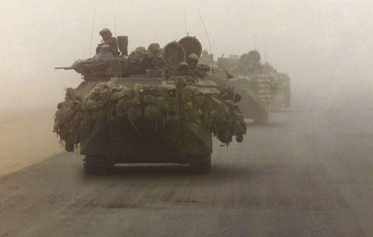 The image depicts military vehicles, specifically tanks, moving through a dusty and foggy environment. The tanks are heavily laden with gear and equipment, indicating they are prepared for deployment or combat. The visibility in the scene is limited due to the dust, which creates a muted atmosphere. Soldiers can be seen perched on top of the tanks, suggesting readiness and vigilance as they navigate the terrain. The overall mood conveys a sense of military operation amid challenging conditions.