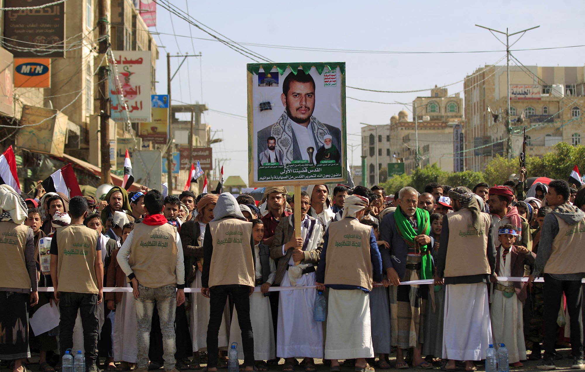 The image depicts a large crowd gathered for a rally or protest in a city, likely in Yemen. In the foreground, a group of people, some wearing beige vests, are standing behind a barricade. They are holding a large poster featuring a portrait of a man, who appears to be a political or religious leader, alongside images of other individuals. The crowd is holding flags, and buildings can be seen in the background, suggesting an urban setting. The atmosphere seems to convey a sense of solidarity or support for the figure on the poster.