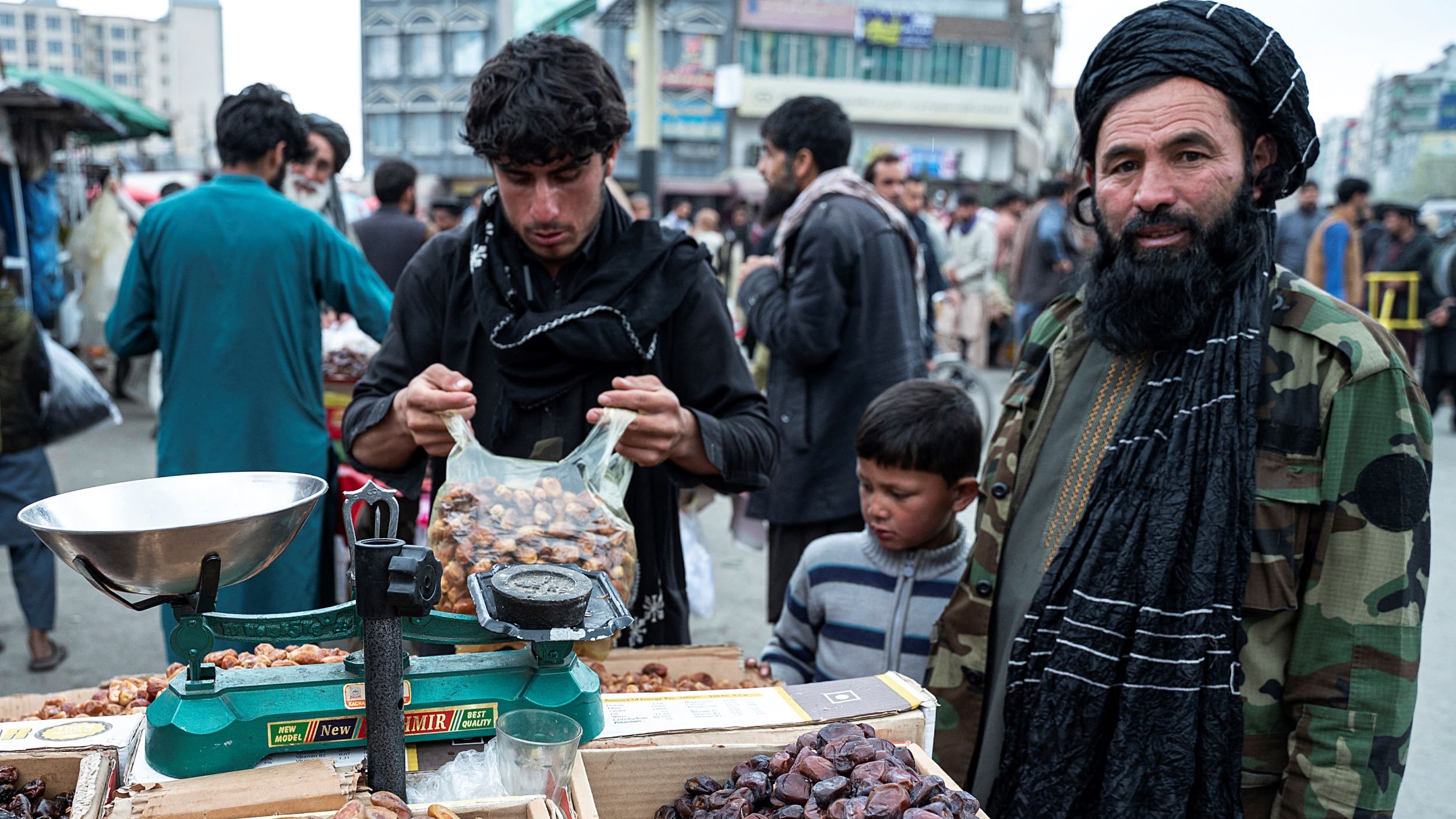 The image depicts a bustling market scene, likely in Afghanistan. In the foreground, there is a vendor displaying various types of dates in baskets and boxes. The vendor, dressed in a traditional outfit and a black turban, stands next to a scale. Another young man is shown packing dates into a bag, while a young boy looks on. The background features a crowd of people, creating a lively atmosphere, with buildings visible in the distance. The overall scene reflects daily life and commerce in a market setting.