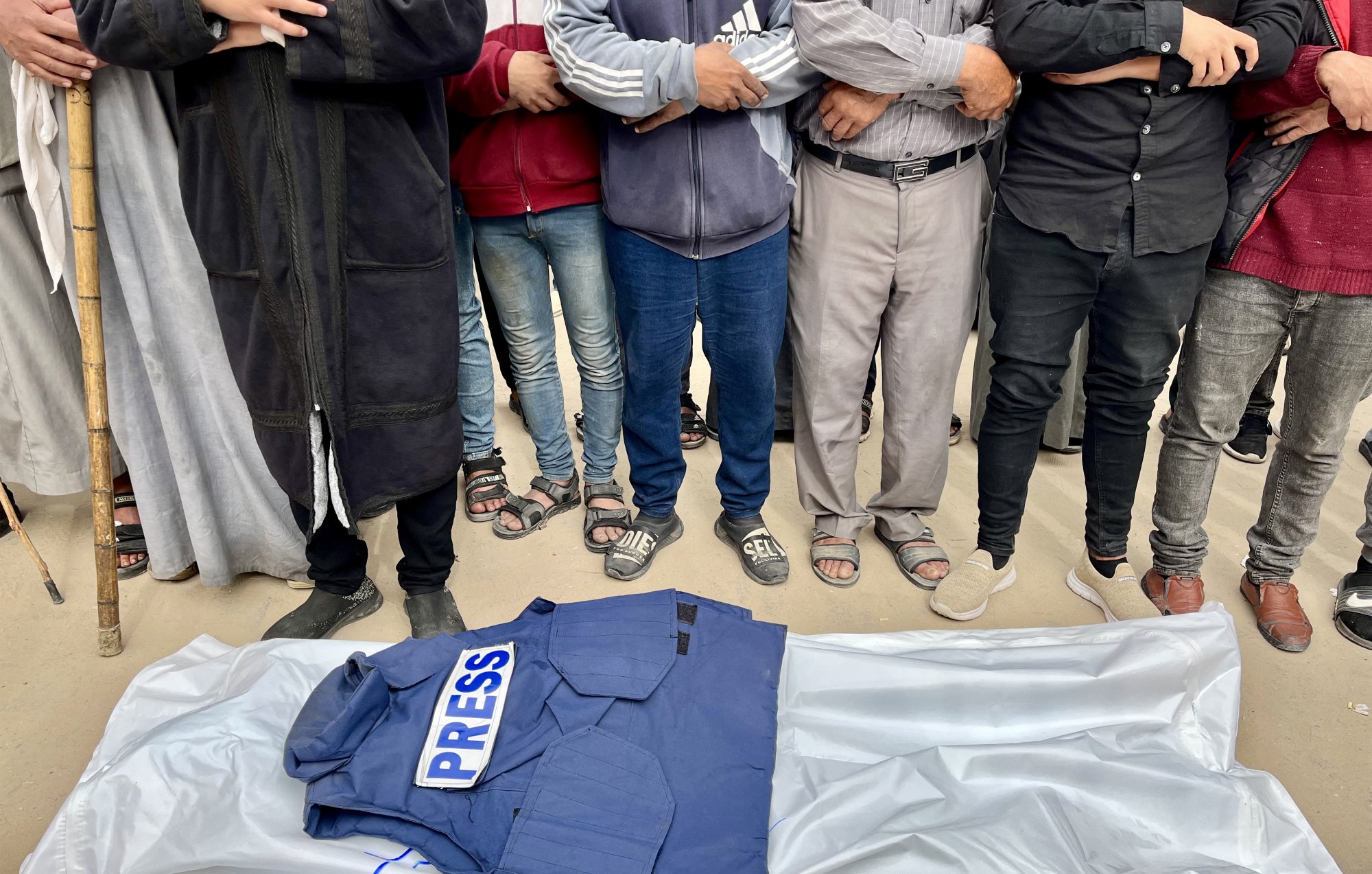 A blue "PRESS" jacket rests on a white sheet, surrounded by people with arms crossed.