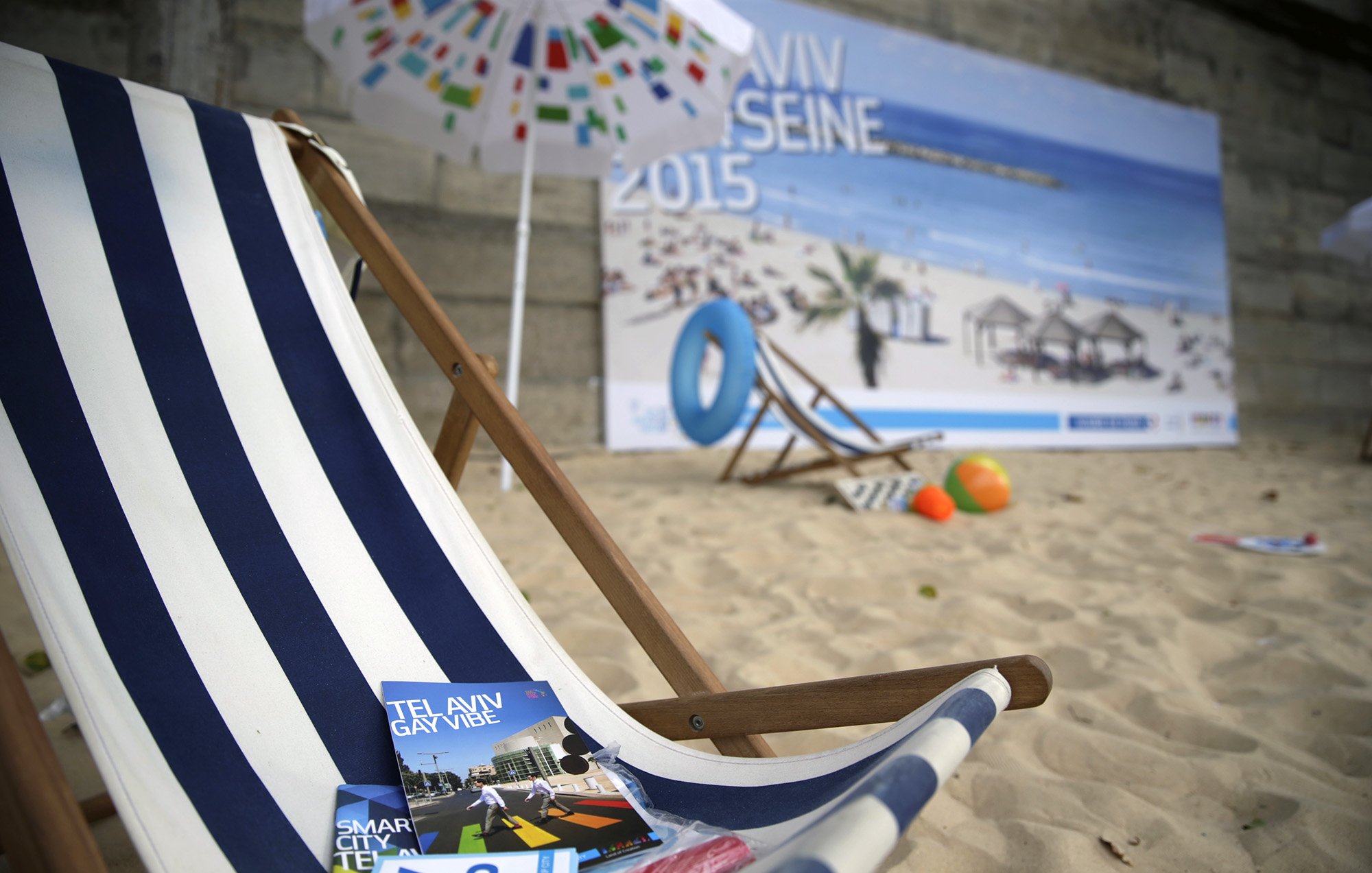 The image features a beach scene with a wooden deck chair adorned with blue and white stripes, set against a backdrop of a sandy area. In front of the chair are brochures for Tel Aviv, including one titled "Tel Aviv Gay Vibe" and another labeled "Smart City Map." The background includes a large poster depicting a sunny beach with umbrellas and people relaxing, suggesting a vibrant, beachside atmosphere.