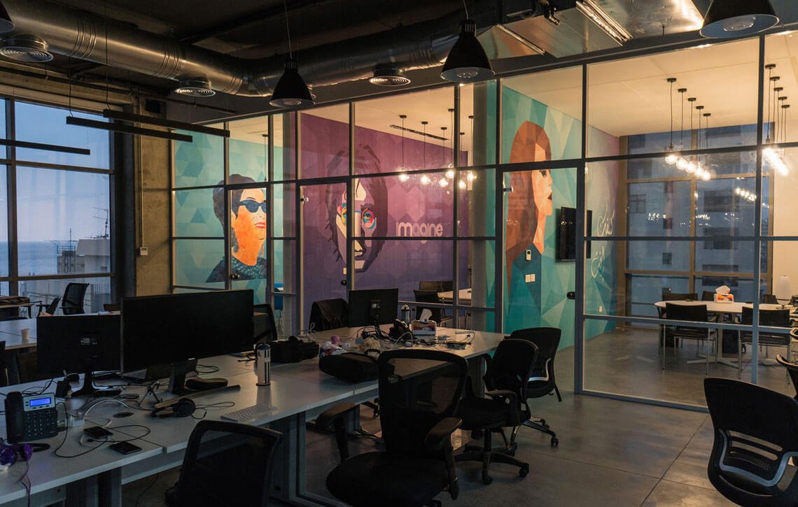 The image shows a modern office space with large glass windows, providing a view of the outside. Inside, there are several workstations with computers and ergonomic chairs. The walls are decorated with vibrant murals featuring stylized portraits of individuals, adding a creative touch to the environment. The overall ambiance is open and contemporary, with industrial elements like exposed ducts and warm lighting fixtures. The space appears to be designed for collaboration and innovation.
