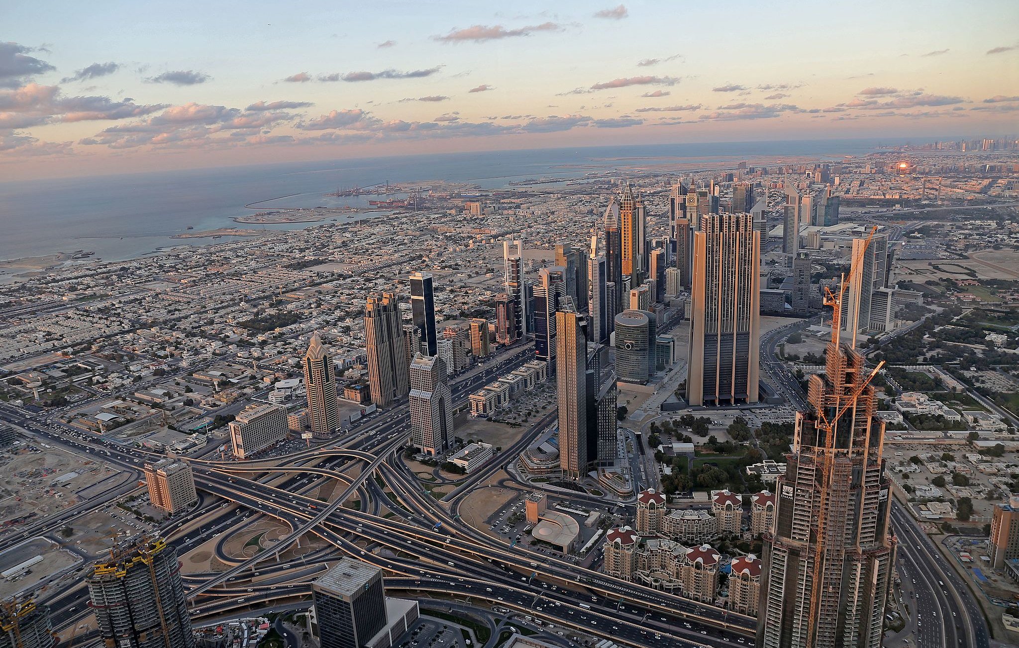 The image depicts a stunning aerial view of a sprawling cityscape, likely a metropolitan area with modern architecture. Skyscrapers dominate the skyline, showcasing a mix of designs and heights. Below, intricate road systems weave through the landscape, indicating a bustling urban environment. In the background, the coastline meets the ocean, with a hint of the horizon showcasing a picturesque sunset. The combination of urban infrastructure and natural beauty creates a striking contrast, highlighting the vibrancy of the city.