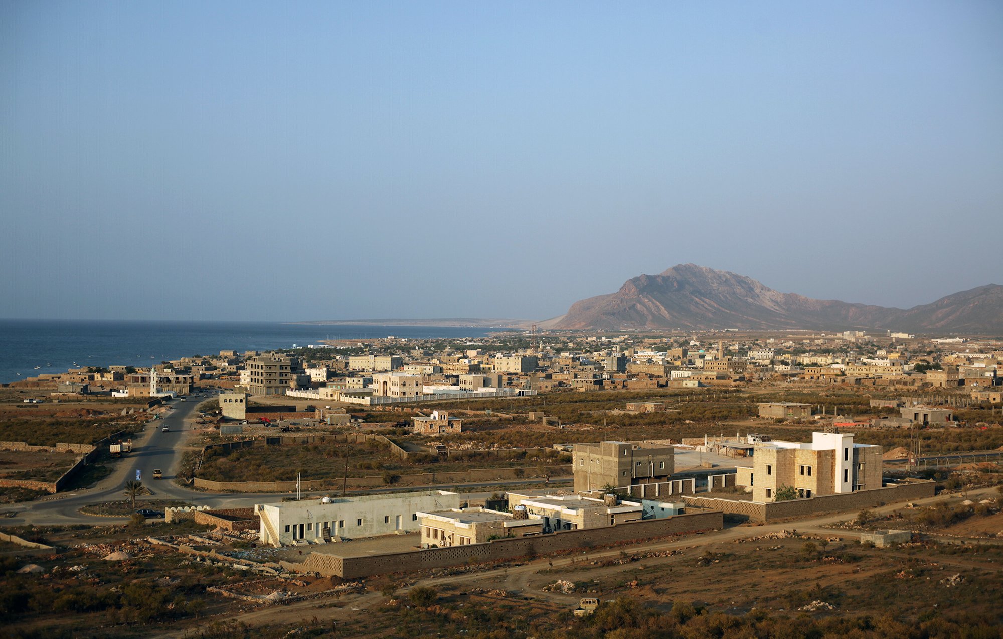 The image depicts a coastal landscape with a view of a small town or village near the ocean. In the foreground, there are a few buildings, some of which are modern, while others appear more traditional. The road winds through the landscape, leading towards the sea. In the background, a mountain rises against a clear sky, adding to the natural beauty of the scene. The overall atmosphere suggests a quiet, possibly rural environment with a mix of natural and man-made elements.