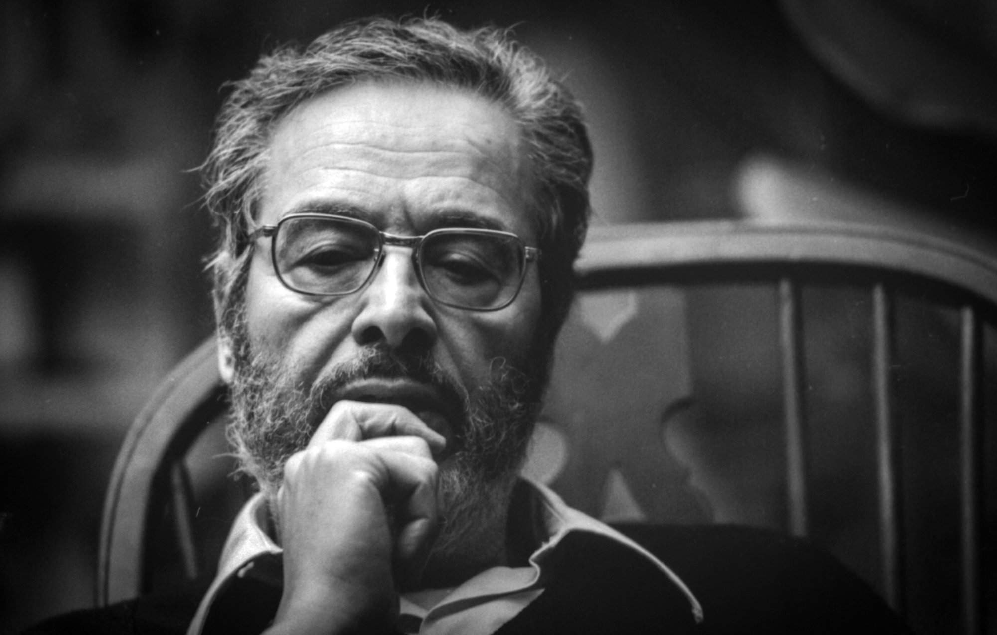 The image features a man with a thoughtful expression, sitting in a wooden chair. He has a beard and glasses, and he appears to be deep in contemplation. The lighting is soft, creating a moody atmosphere, and the background is blurred, focusing attention on his contemplative demeanor. The photograph has a classic black-and-white aesthetic, emphasizing the seriousness of the moment.