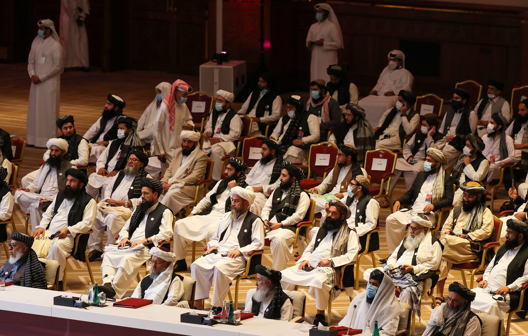 The image depicts a large gathering of individuals seated in a formal setting, likely during a conference or meeting. The attendees are predominantly dressed in traditional attire, including long robes and turbans. Some individuals are wearing face masks, suggesting a concern for health or safety. The arrangement indicates a structured seating plan, with a few individuals at the front possibly in leadership roles. The venue appears to be elegant, with ornate decorations and a spacious layout typical of an official or ceremonial event.