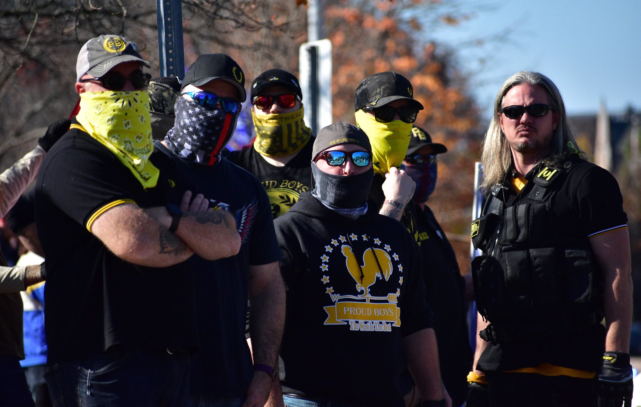 The image depicts a group of people standing together, wearing clothing that features dark colors and some yellow accents. Many of them have their faces covered with bandanas or masks, and they are wearing sunglasses. Their expressions and postures suggest a sense of solidarity or confrontation. The group is likely gathered for a specific event or rally, as indicated by their coordinated appearance. In the background, there are trees and other people visible, suggesting an outdoor setting.