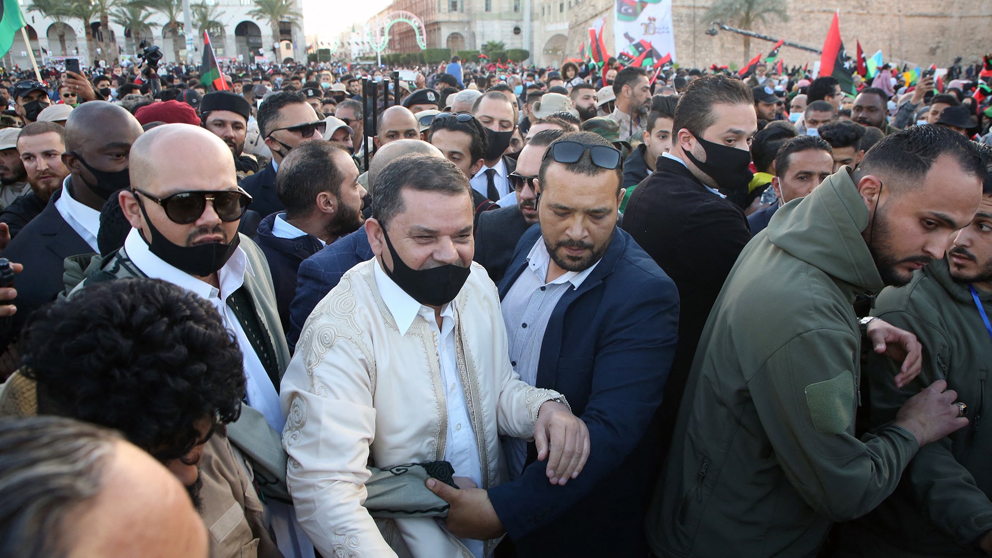 The image depicts a crowd of people gathered at an outdoor event, likely a political or social gathering. In the foreground, a man wearing a white traditional outfit is surrounded by a group of security personnel or supporters. Many individuals in the crowd are wearing black face masks. The background features various flags and buildings, indicating a festive or significant atmosphere. The scene appears to be lively, with a sense of excitement among the gathered individuals.
