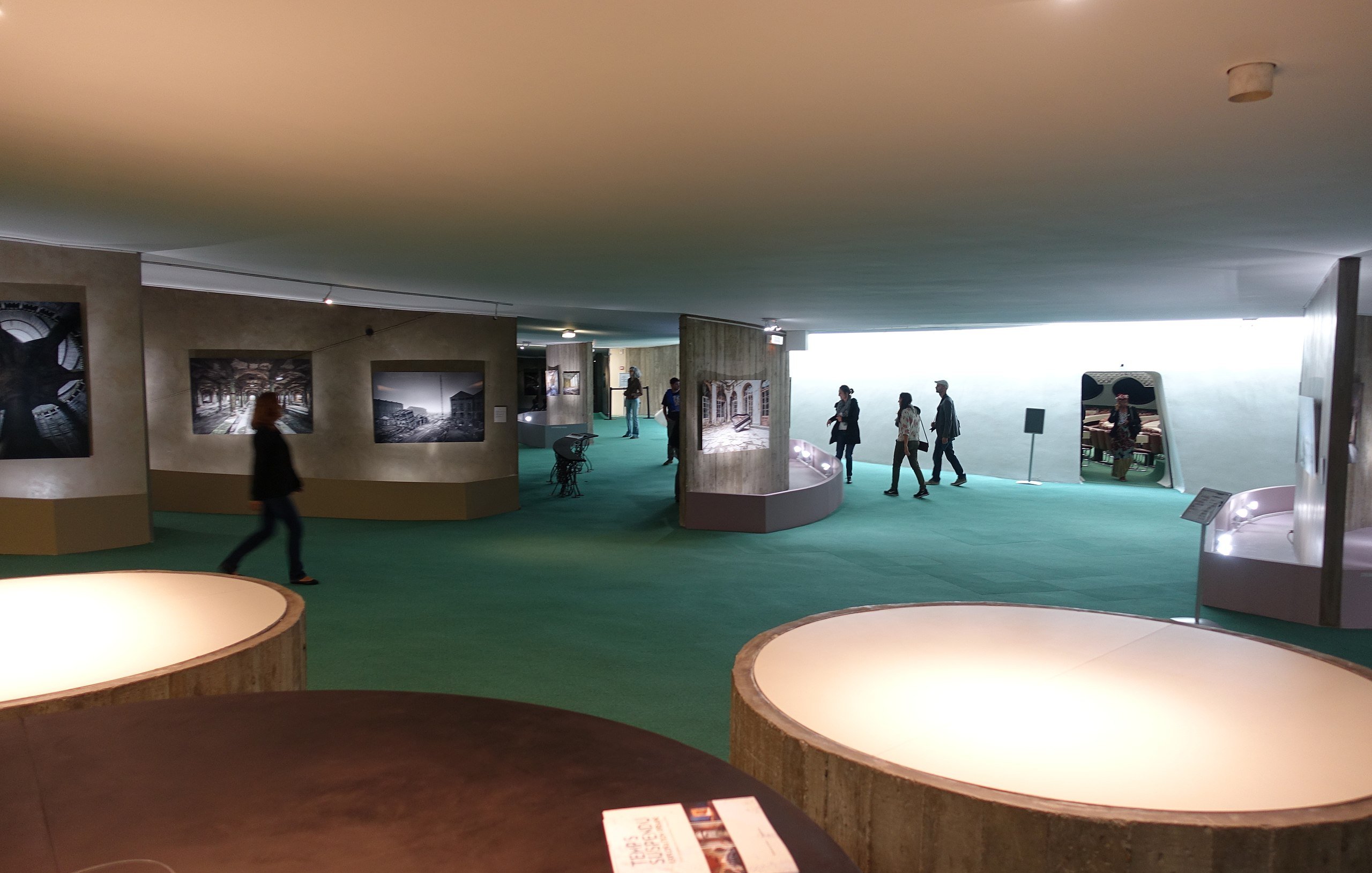 The image depicts an interior space, likely an art gallery or museum. The floor is covered with green carpeting, and the walls display a series of black-and-white photographs. Several people are walking through the space, engaged with the exhibits. There are curved display structures that showcase the photographs. The lighting appears bright and modern, creating an inviting atmosphere.