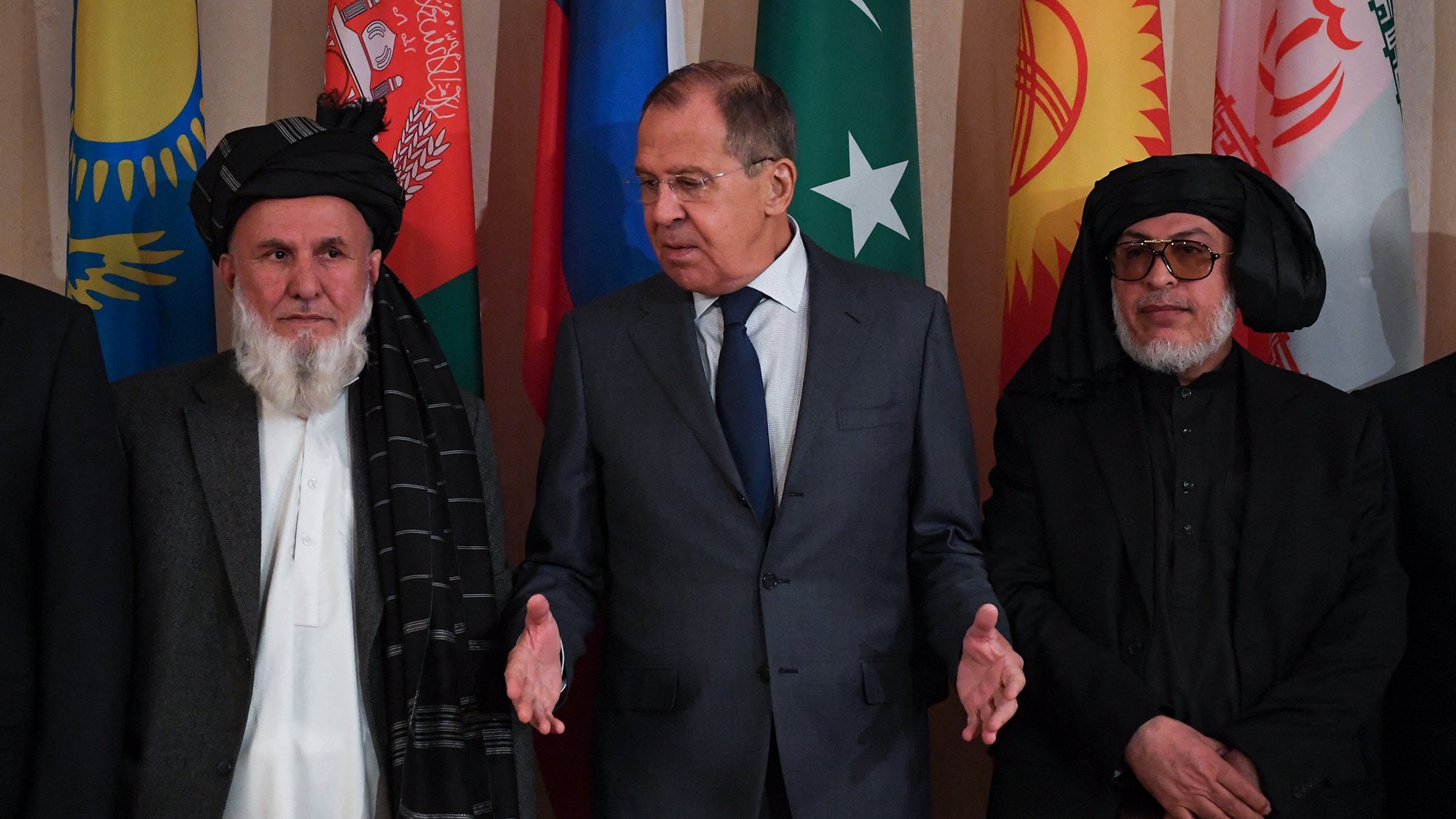The image features a group of men standing in front of a backdrop adorned with various national flags. In the center is a man in a suit, possibly a diplomat or official, gesturing as he speaks. To his left and right are two men dressed in traditional attire, including a shalwar kameez and a shawl. The overall atmosphere suggests a formal meeting or diplomatic discussion, likely concerning regional issues or cooperation.