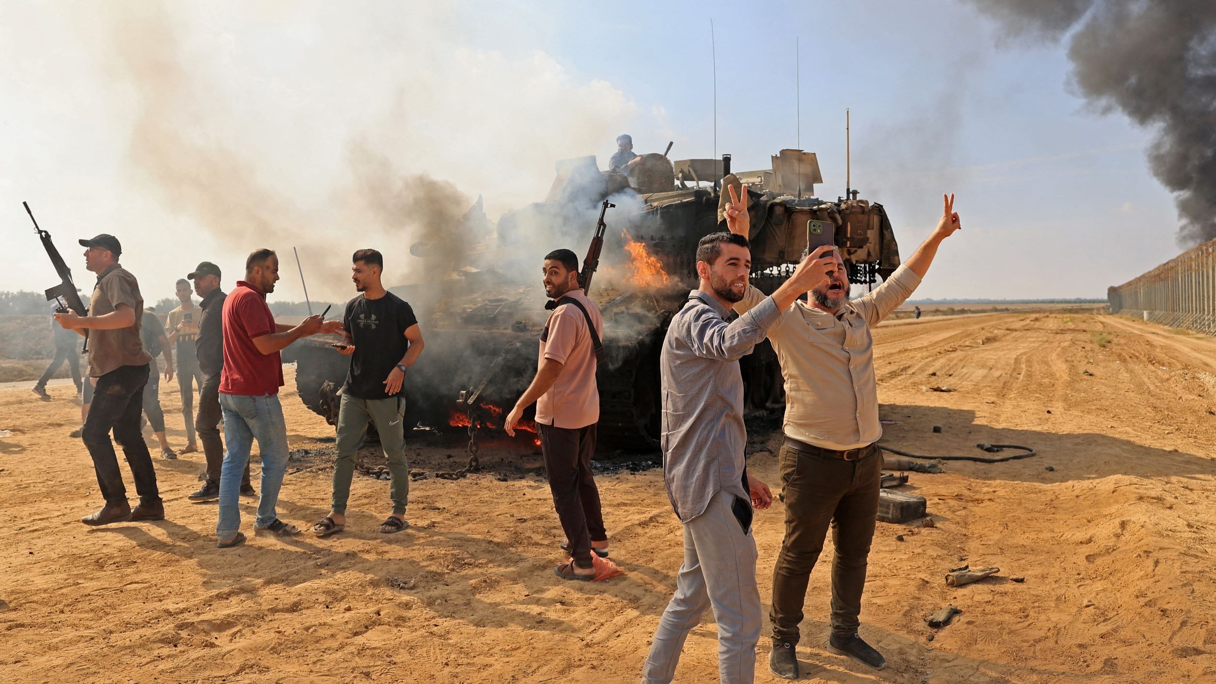 The image depicts a chaotic scene in a sandy area where a group of individuals appears to be celebrating or protesting near a burning military vehicle. Smoke rises from the vehicle, creating a dramatic backdrop. Some people are visible holding firearms, while others are raising their hands, possibly in a gesture of victory. The environment suggests a conflict or demonstration, with a tense atmosphere reinforced by the presence of smoke and the military vehicle. The landscape is open, with a barrier visible in the background, indicating a border or contested area.