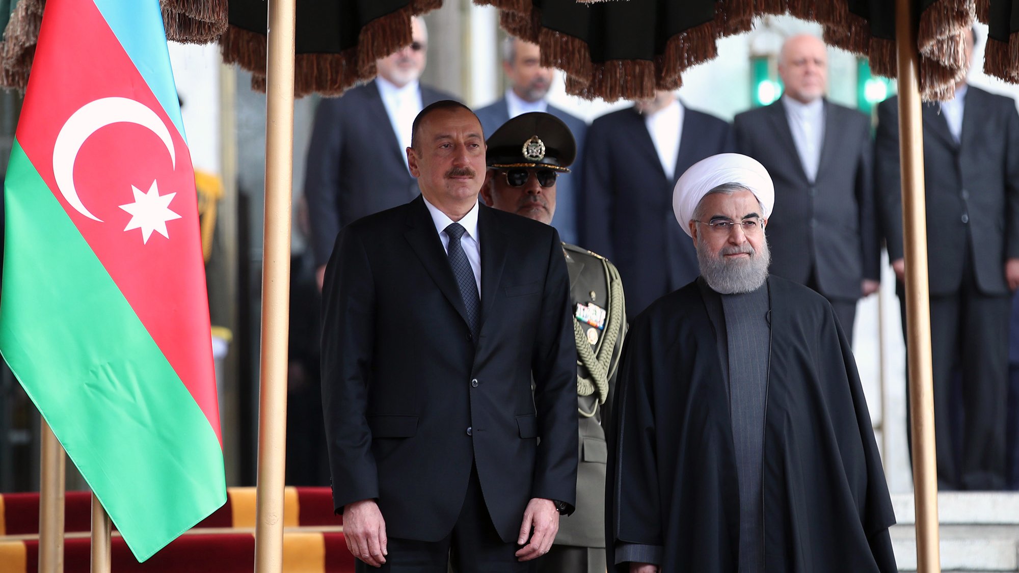 The image depicts a formal meeting between two leaders, presumably from Azerbaijan and Iran, standing side by side. In the foreground, one leader is dressed in a suit, while the other is wearing a dark robe and headpiece. Behind them, a group of individuals can be seen, and a large flag of Azerbaijan is displayed prominently on the left. The setting appears to be an official location, likely a government building, with stairs and a red carpet visible.
