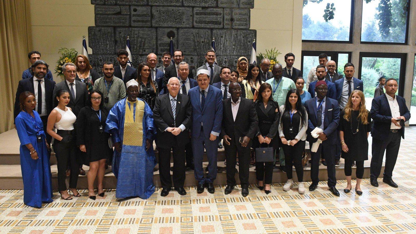 The image shows a large group of people gathered together for a formal event. They are standing in a room with a decorative backdrop that includes flags and a large, textured wall. The group consists of men and women of diverse backgrounds, and many are dressed in professional attire. Some individuals wear traditional cultural clothing. Everyone appears to be smiling, and they are posed together for a photograph, likely to commemorate the occasion. The setting is bright and well-lit, contributing to a positive atmosphere.