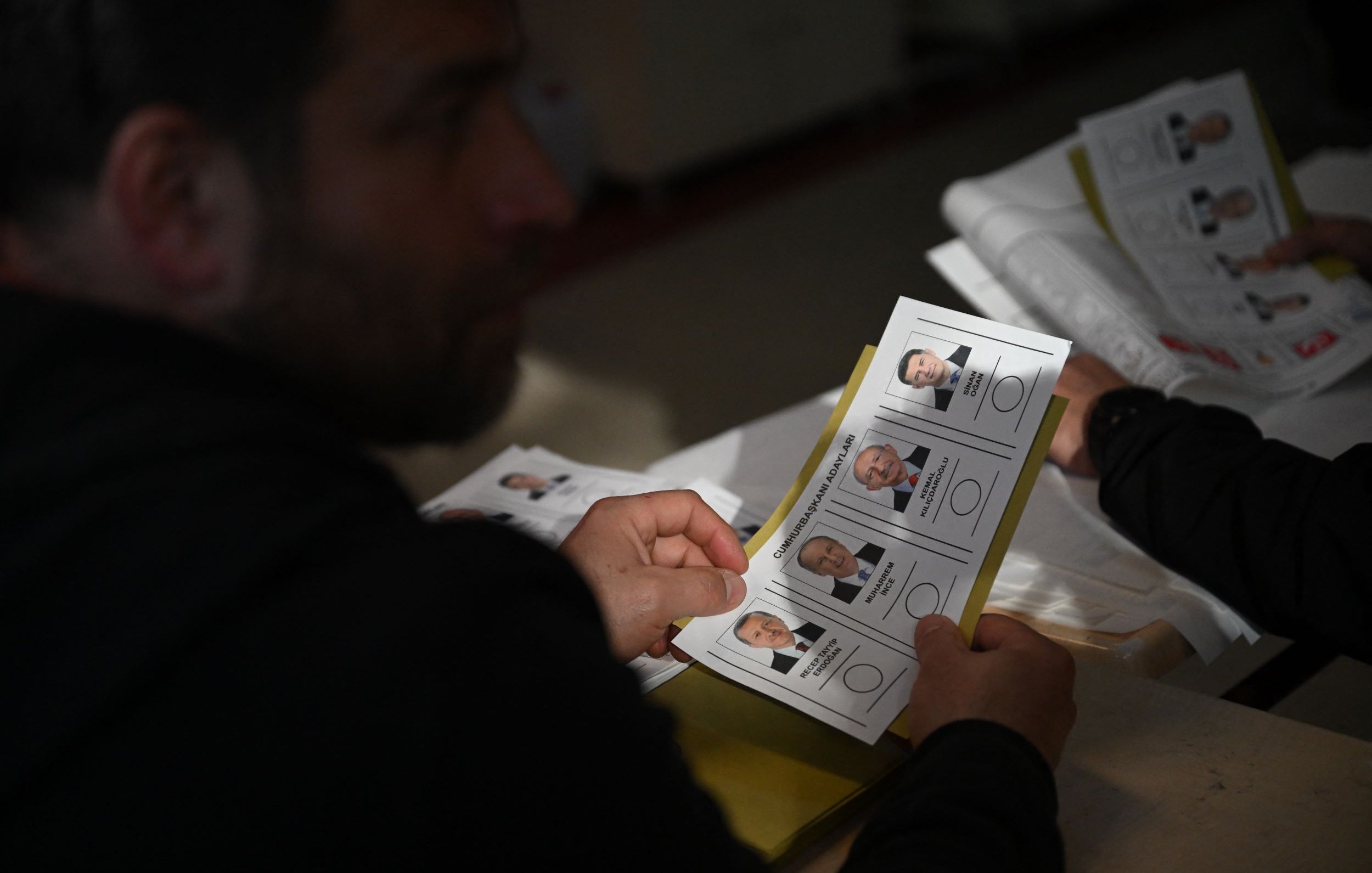 The image shows a person, partially visible, holding a ballot or voting card that features photographs and names of candidates. The setting appears to be a polling station, with multiple ballots or documents visible. The individual seems to be examining the options before making a decision. The lighting suggests an indoor environment, likely during a voting process.
