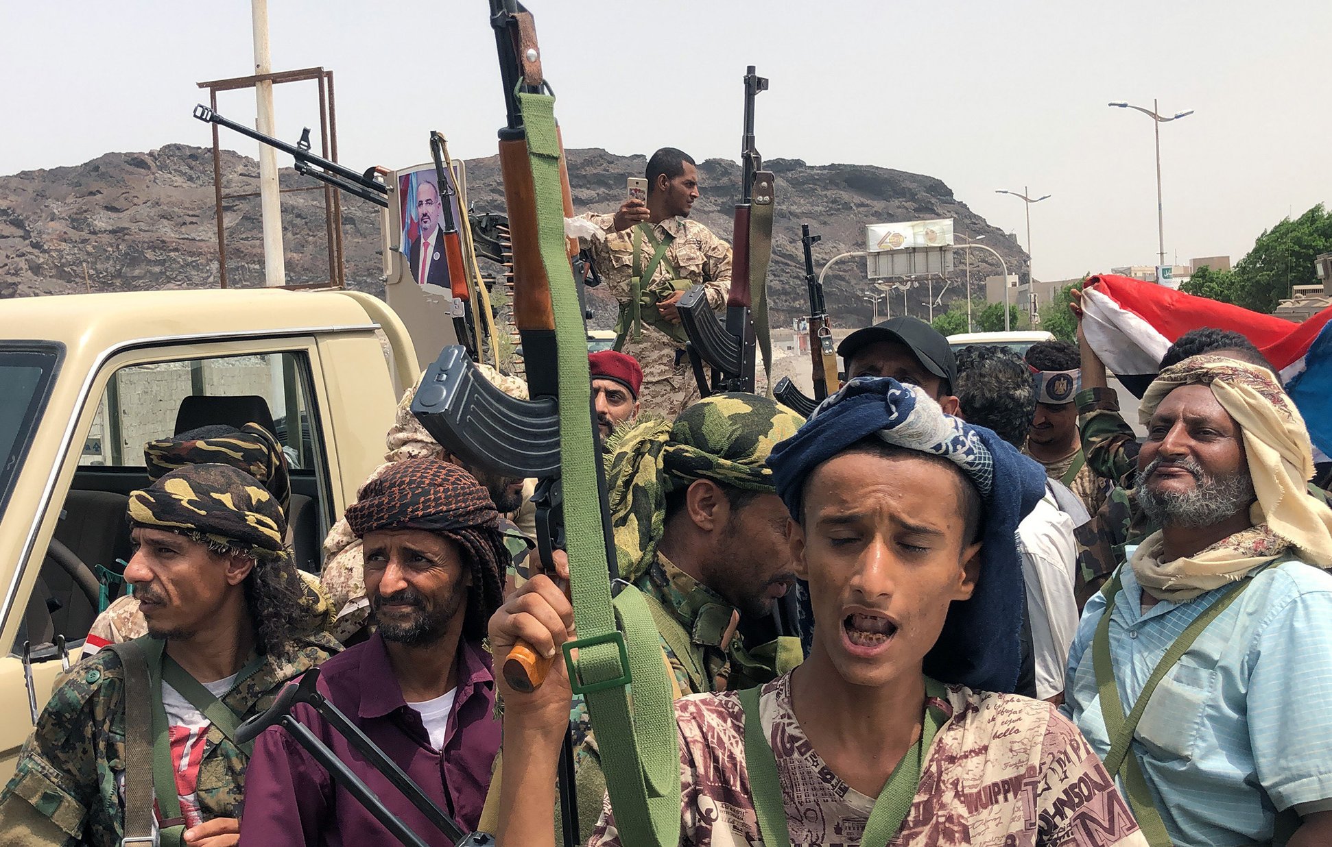 The image depicts a group of armed individuals standing together in an outdoor setting. They are dressed in military-style clothing and some wear traditional headscarves. Several of them are holding rifles and appear to be engaged in a rally or demonstration. The background features a vehicle and a large rock formation, with a hint of a cityscape. A flag is visible, adding to the atmosphere of the gathering, which seems to pertain to a political or military context. The expressions and postures of the individuals reflect a sense of determination or unity.
