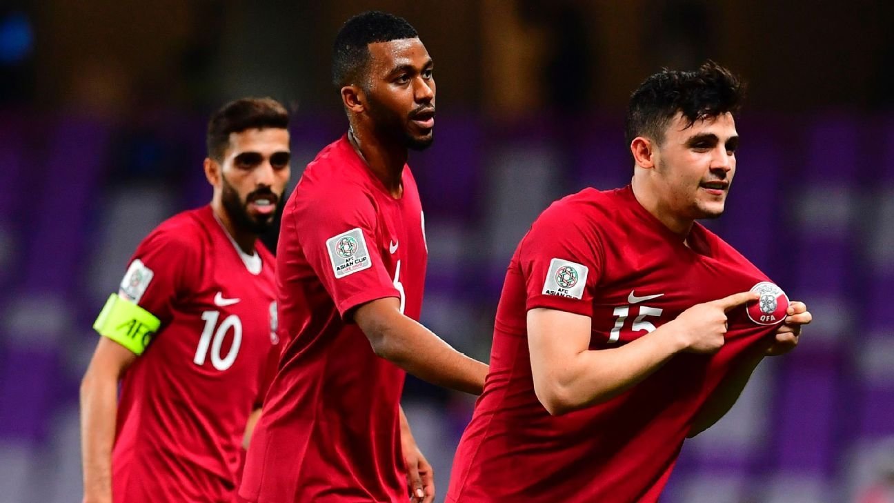 The image captures a moment from a football (soccer) match featuring players in red jerseys representing Qatar. One player, wearing the number 15, is celebrating and pulling his jersey up, possibly revealing a symbol or crest underneath. The player in the foreground seems excited and is likely celebrating a goal. Behind him, two other teammates are visible; one is wearing the number 10 and the other appears to be the team captain, as indicated by the captain's armband on his arm. The stadium setting suggests a significant match, likely during a tournament.