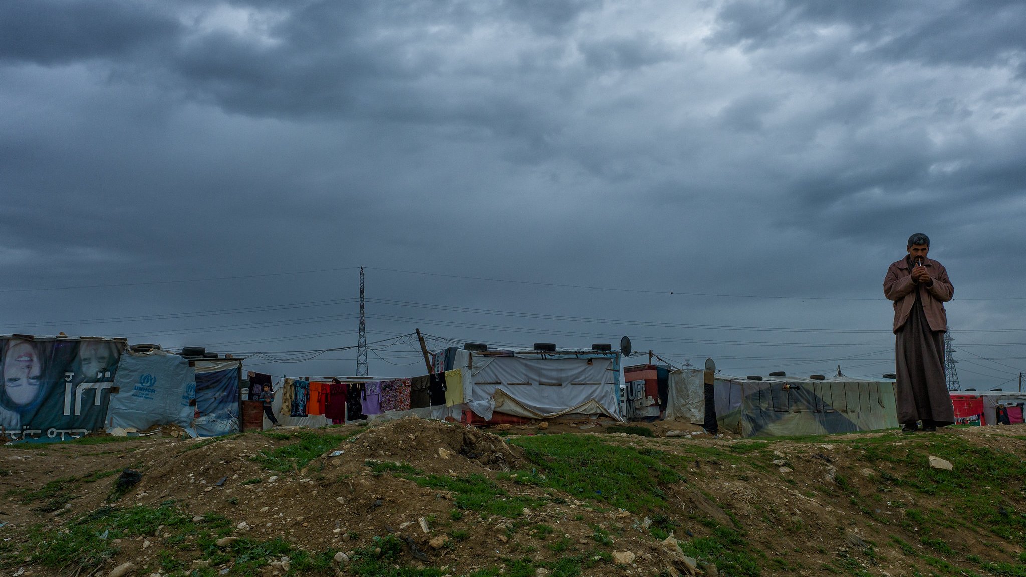 The image depicts a somber landscape under a cloudy sky. In the foreground, a lone figure stands, appearing pensive or contemplative. The background features makeshift structures, likely tents or shanties, indicating a temporary or informal settlement. These structures are adorned with various colors and materials, reflecting a harsh living environment. The overall atmosphere conveys a sense of solitude and resilience against the backdrop of an overcast sky.