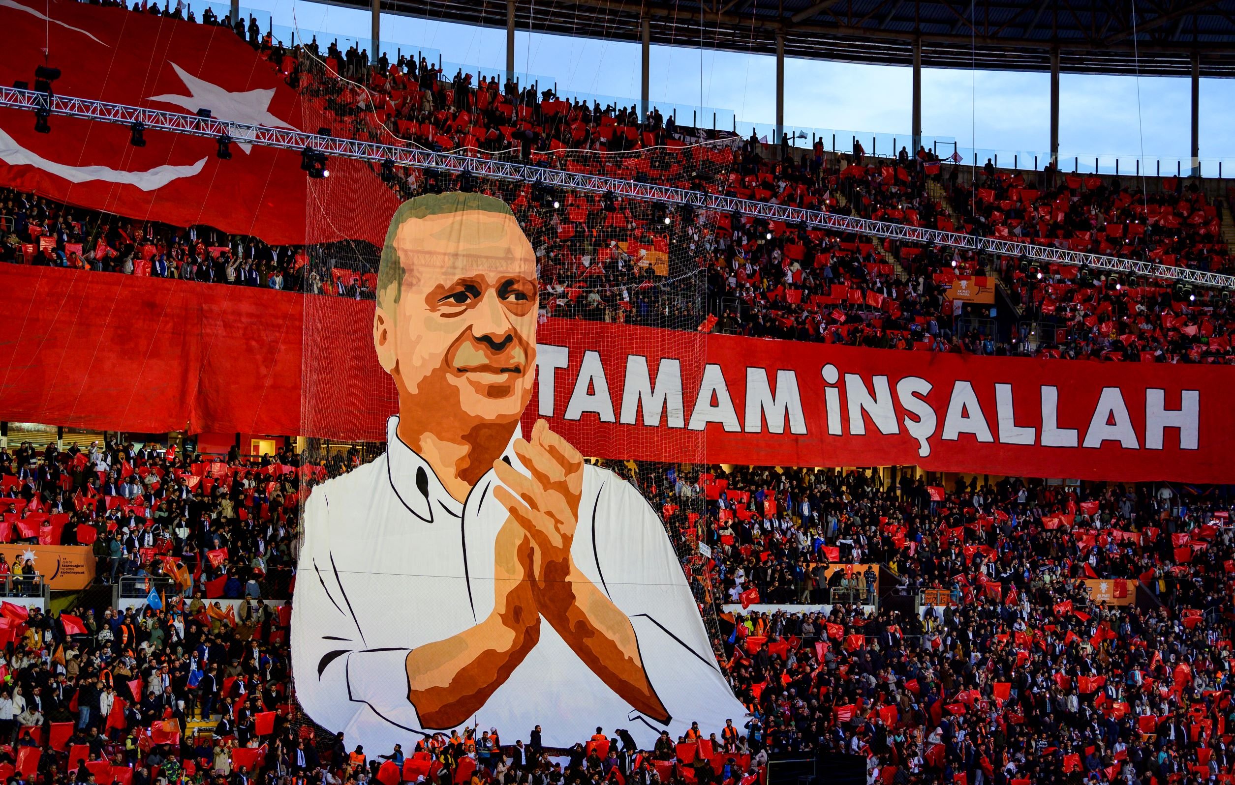 The image depicts a large crowd in a stadium, with a significant banner displayed prominently. The banner features a representation of a person, possibly a political figure, who is shown applauding. Surrounding the figure are enthusiastic supporters holding a sea of red flags or banners. The text "TAMAM İNŞALLAH" is written in bold letters, which translates to "It will be done, God willing" in English, conveying a message of hope or determination. The overall atmosphere appears vibrant and charged with energy, indicative of a political rally or celebration.